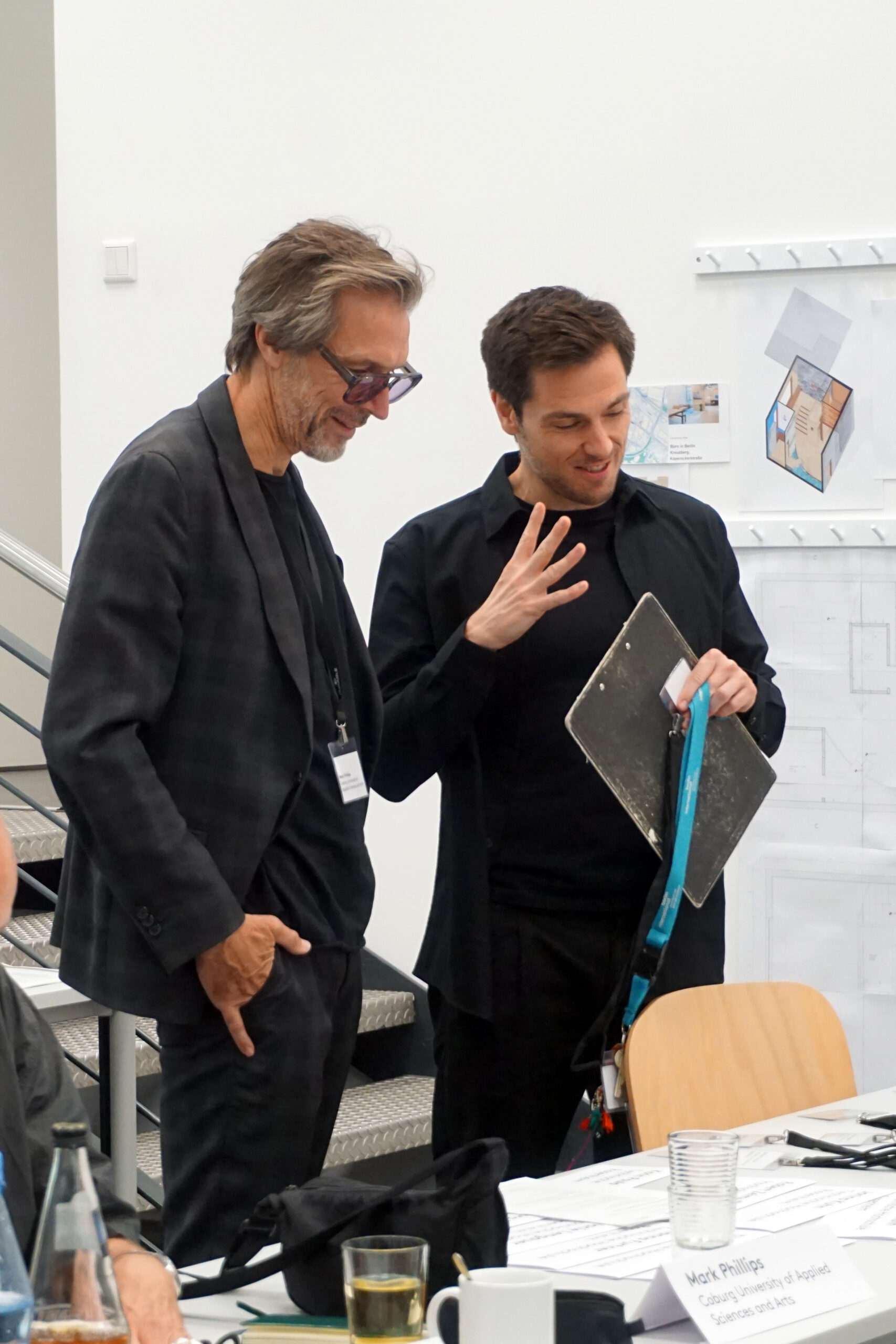 Two men are standing by a table covered with papers and office supplies at Hochschule Coburg. One is holding a clipboard and gesturing. Engaged in conversation, they smile warmly. In the background, a staircase and a wall adorned with blueprints reflect their academic environment.