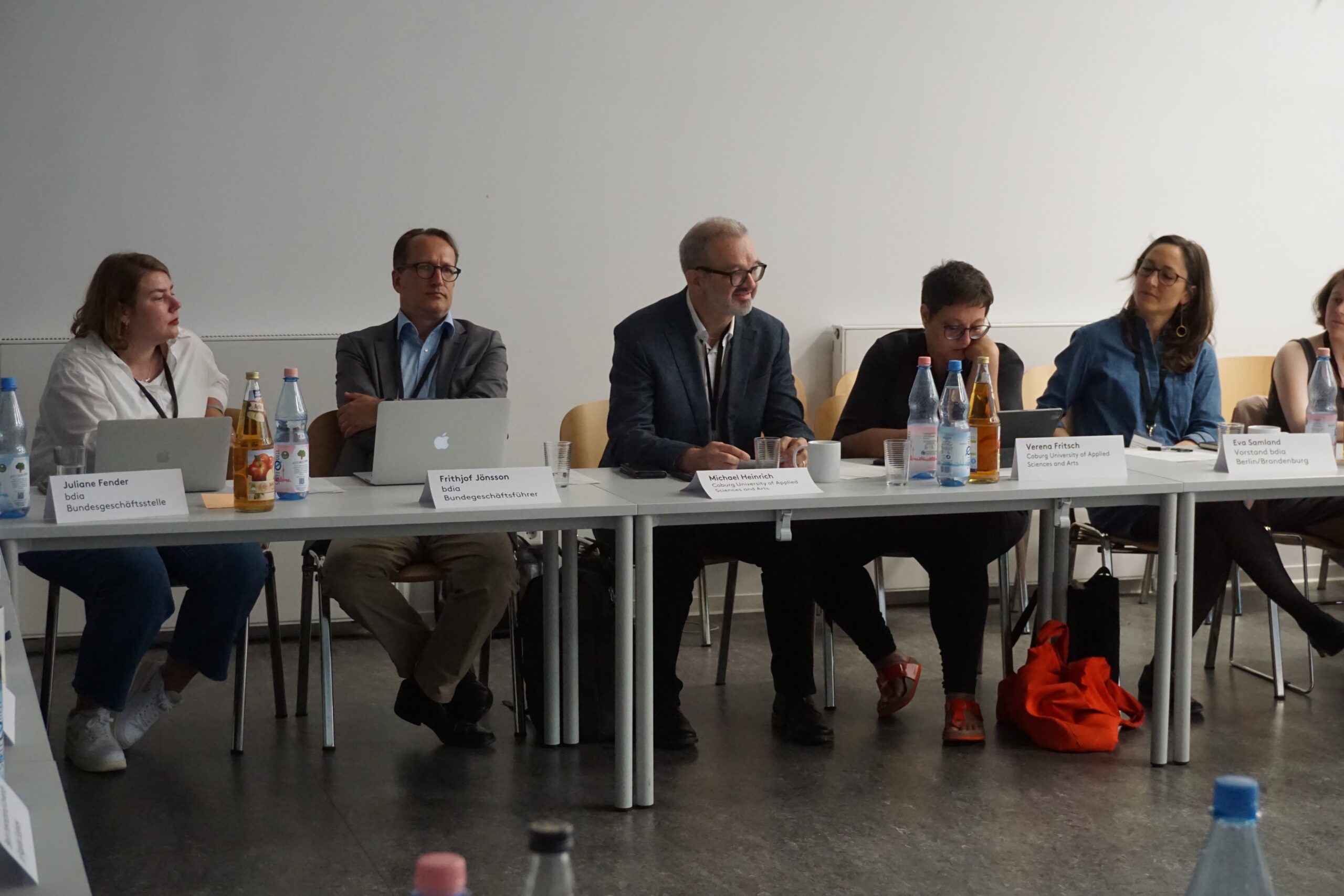 A group of six people sit at a long white table, participating in a panel discussion at Hochschule Coburg. Some have laptops open, and several bottled drinks are on the table. They appear engaged, with nameplates in front of each person.