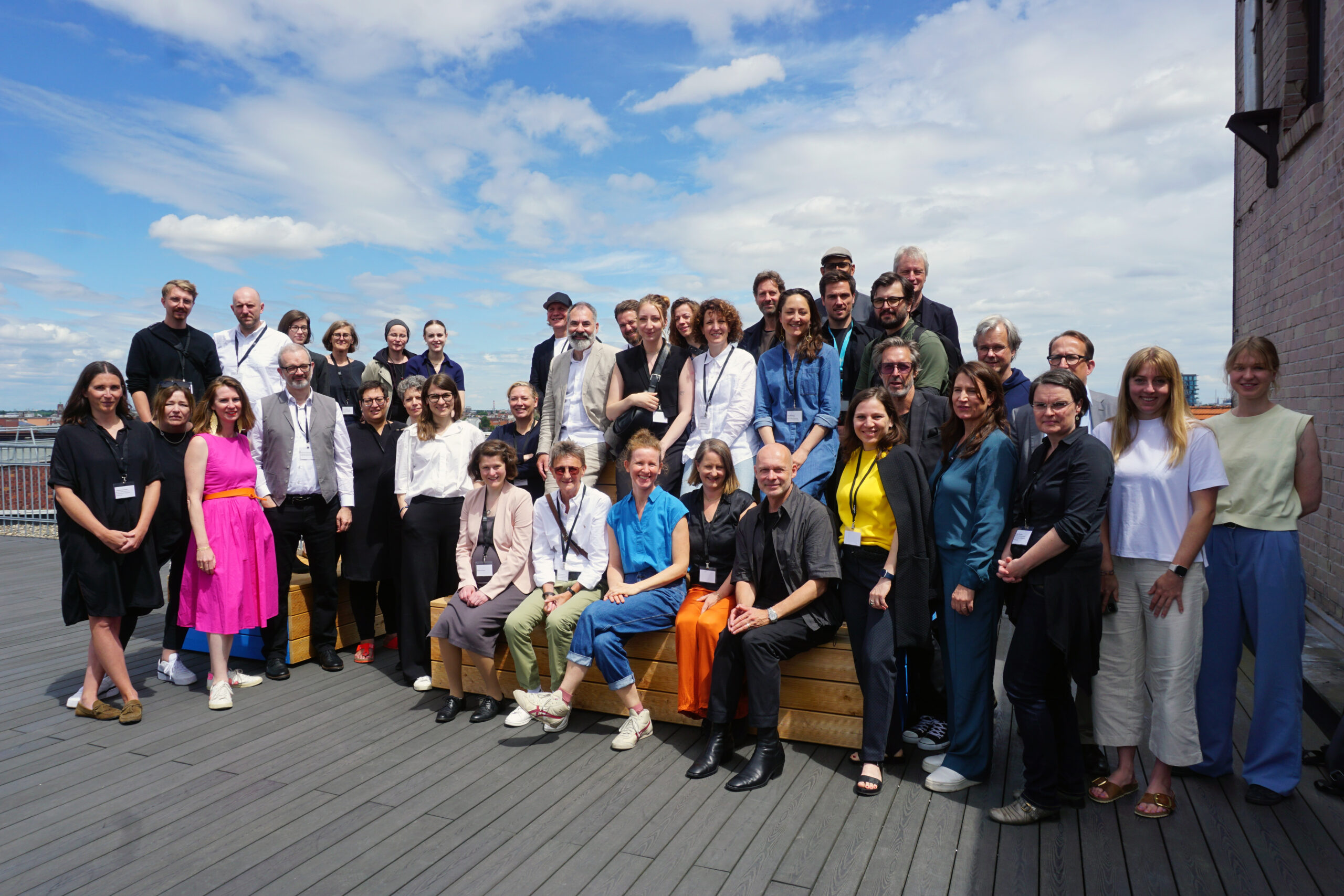 A diverse group of about 30 people, dressed in various casual and professional outfits, pose together on a wooden deck under the clear blue sky. Some are seated on a bench while others stand behind them, embodying the vibrant community of Hochschule Coburg.