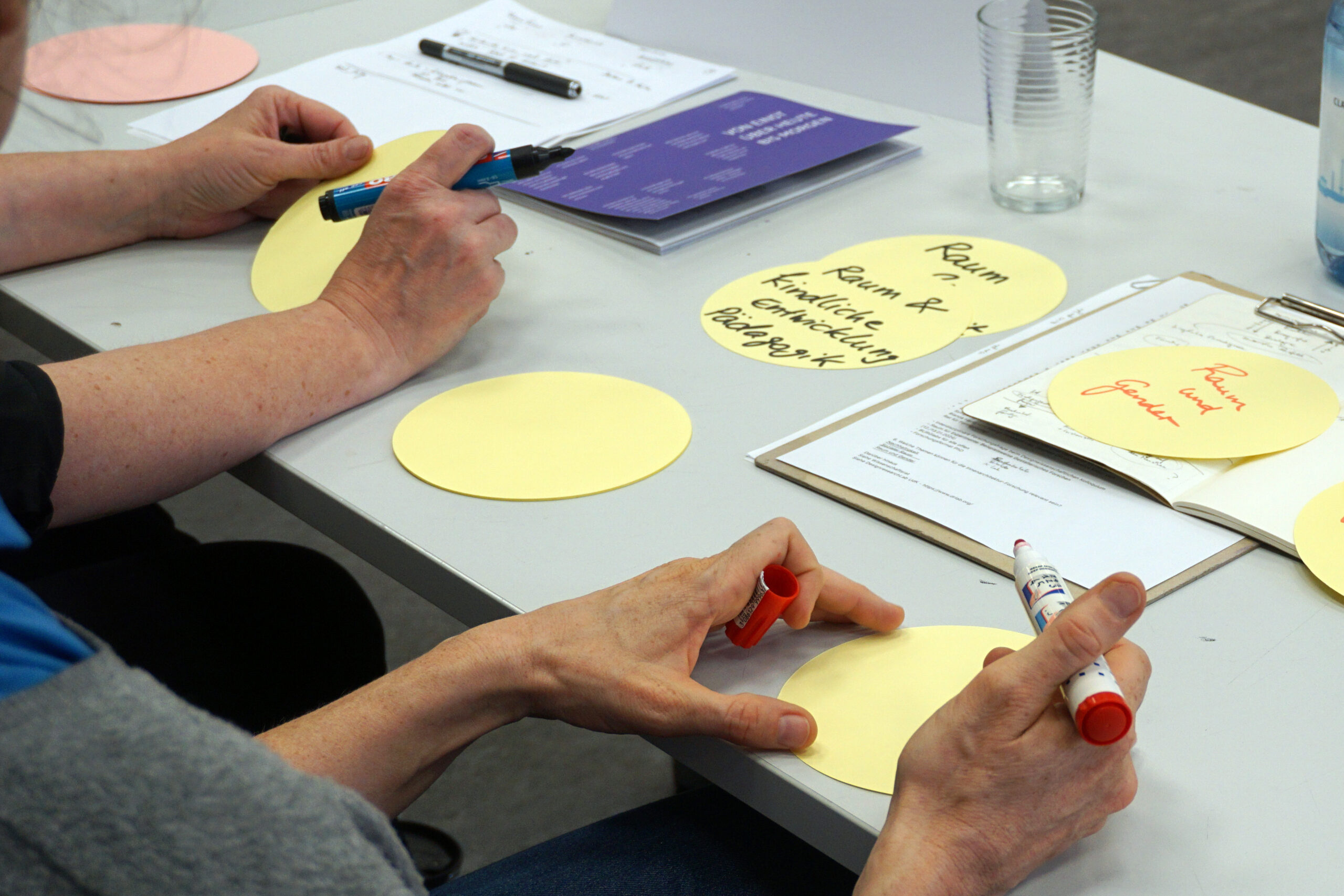 At a Hochschule Coburg desk, two people write with markers on yellow paper circles. One wields a red pen, the other a blue pen, as notes and a purple booklet lie nearby. A transparent cup and bottle complete the academic scene.