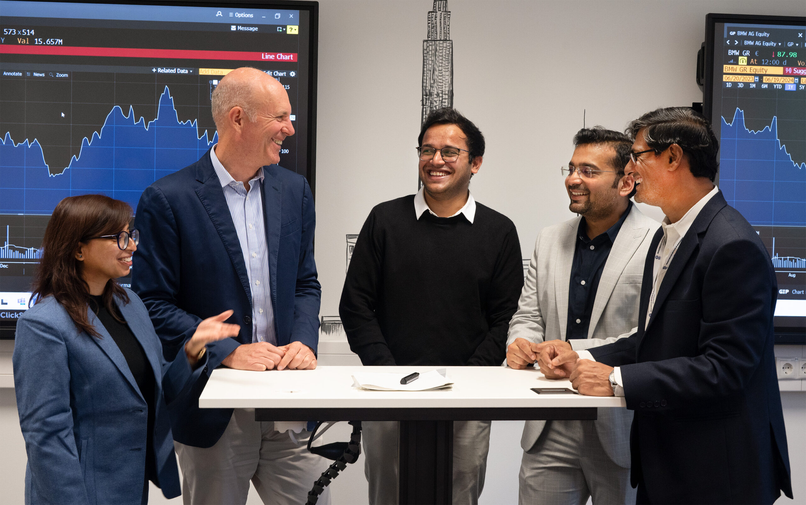 A group of five professionals, three men and two women, are standing around a table at Hochschule Coburg, smiling and engaging in conversation. Behind them, two screens display financial graphs and charts in an office setting.