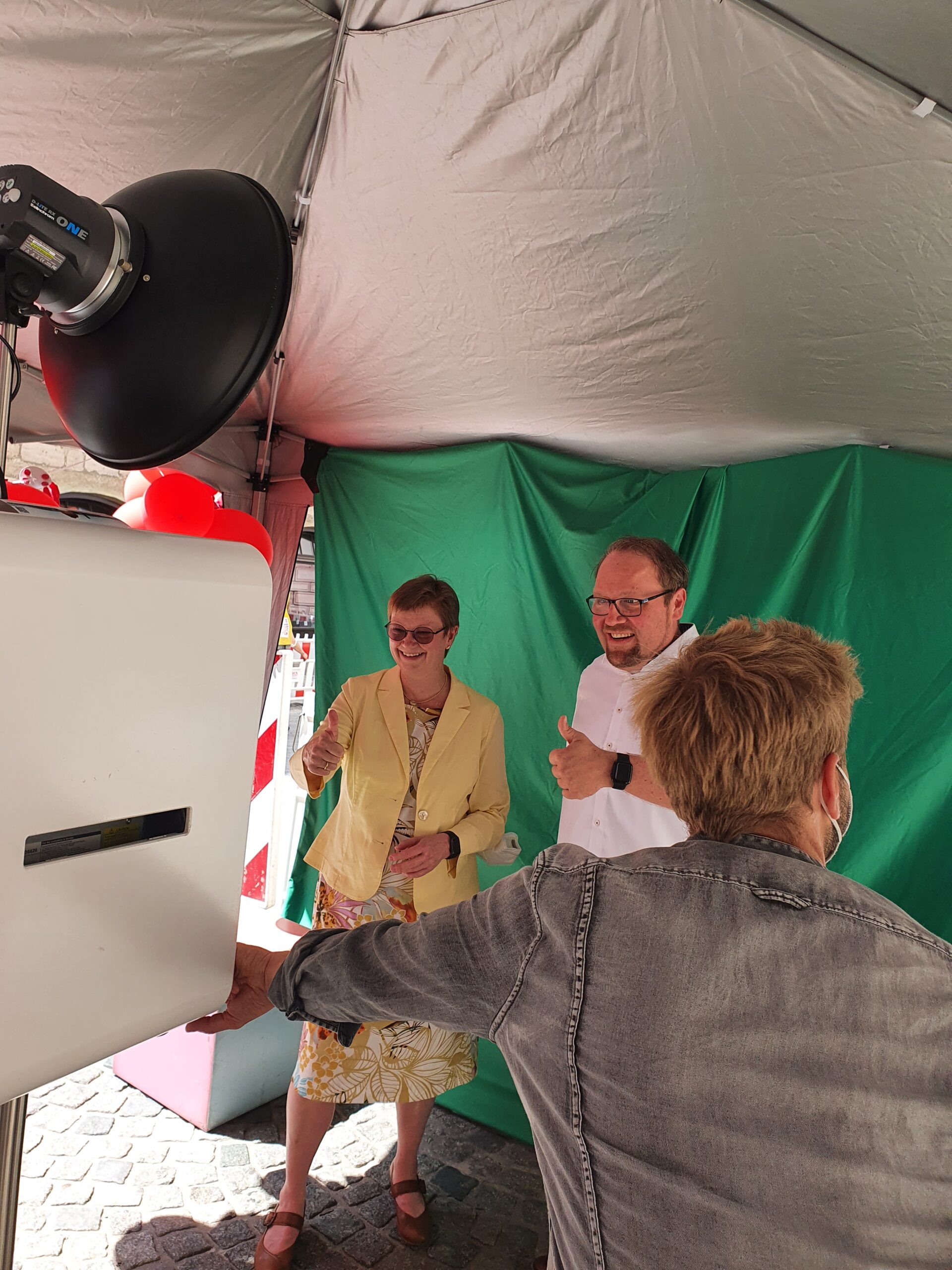 Drei Personen posieren in einer Fotokabine mit Greenscreen-Hintergrund unter einem Baldachin an der Hochschule Coburg. Zwei von ihnen, links, strecken den Daumen nach oben. Im Vordergrund sind eine Kamera und ein großes Licht zu sehen, umgeben von festlicher Dekoration.