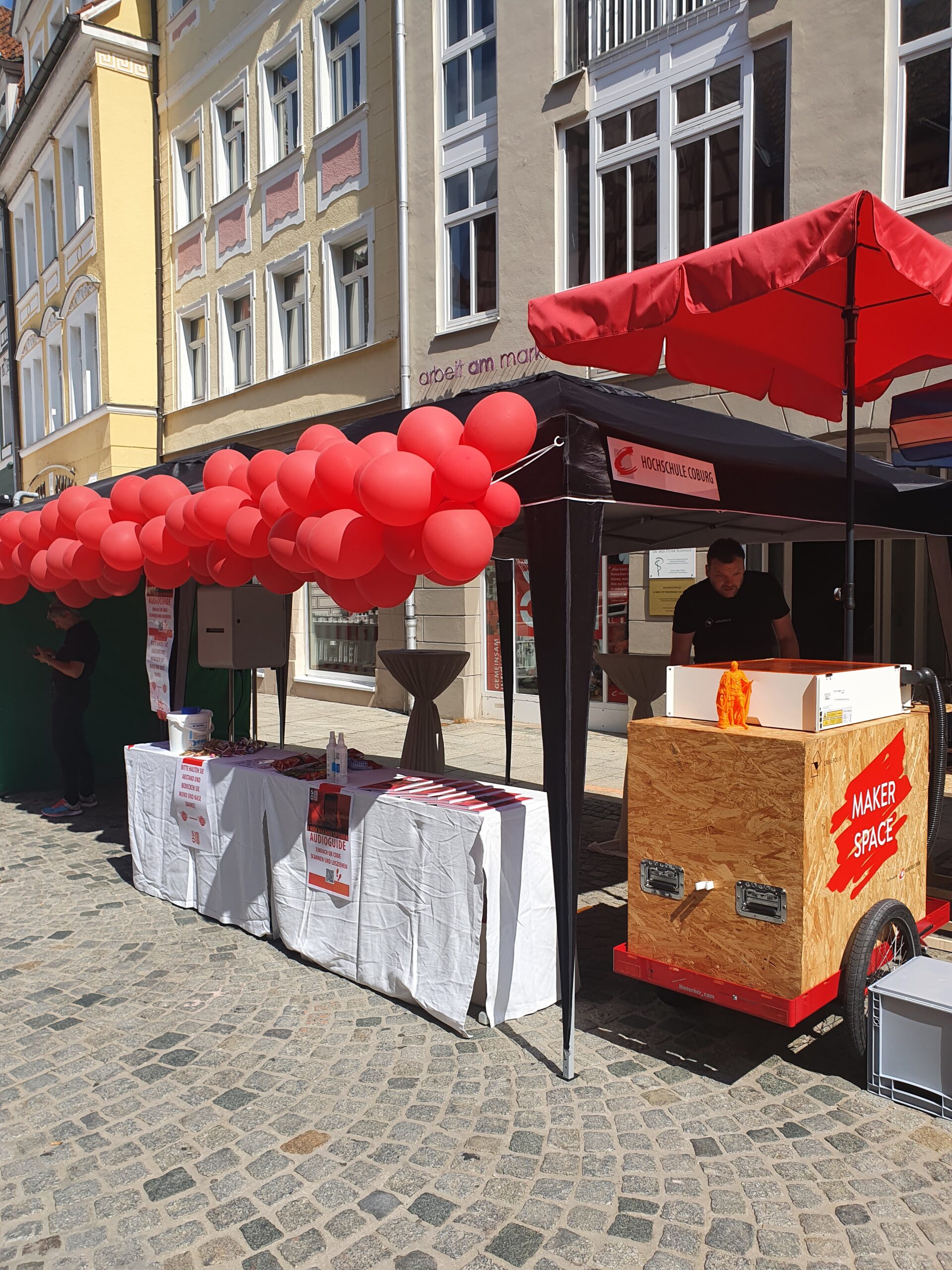 Eine Straßenszene zeigt einen Stand mit einem roten Vordach und einem Wagen mit der Aufschrift „Maker Space“. Rote Luftballons schmücken das Vordach. Tische mit Flyern der Hochschule Coburg sind aufgestellt, eine Person steht hinter dem Tresen. In der Nähe runden bunte Gebäudefassaden die lebendige Atmosphäre ab.