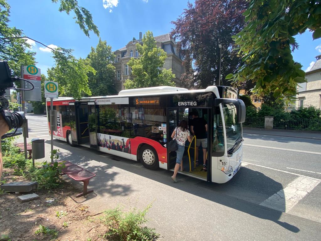 An einem sonnigen Tag steigt eine Person in einen rot-weißen Stadtbus mit der Aufschrift „Einstieg“ in der Nähe der Hochschule Coburg. Die Straße ist von Bäumen und älteren Gebäuden gesäumt. An der Bushaltestelle steht eine Bank.