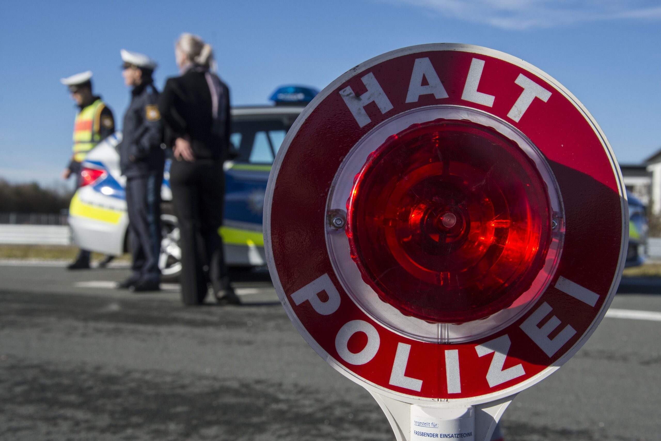 Im Fokus steht ein Stoppschild der deutschen Polizei mit der Aufschrift „Halt Polizei“. Im Hintergrund stehen zwei uniformierte Beamte und eine Frau neben einem Polizeiauto mit dem Logo der Hochschule Coburg auf einer Straße. Der Himmel ist klar und blau.