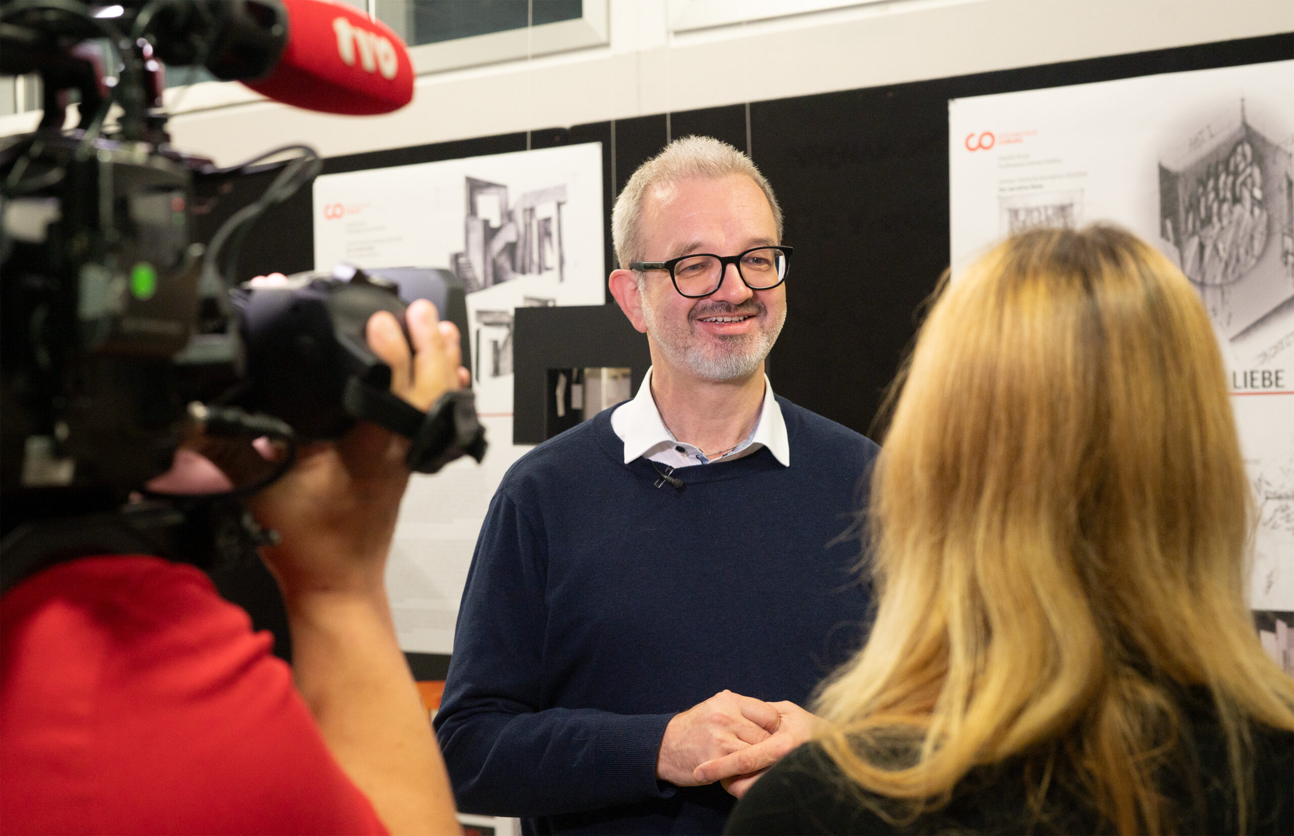 A man with glasses and a beard is being interviewed by a TV crew at Hochschule Coburg. Smiling, he wears a navy sweater over a white shirt. A camera and microphone are visible, while a woman with blonde hair stands with her back to the camera. Posters decorate the wall behind them.