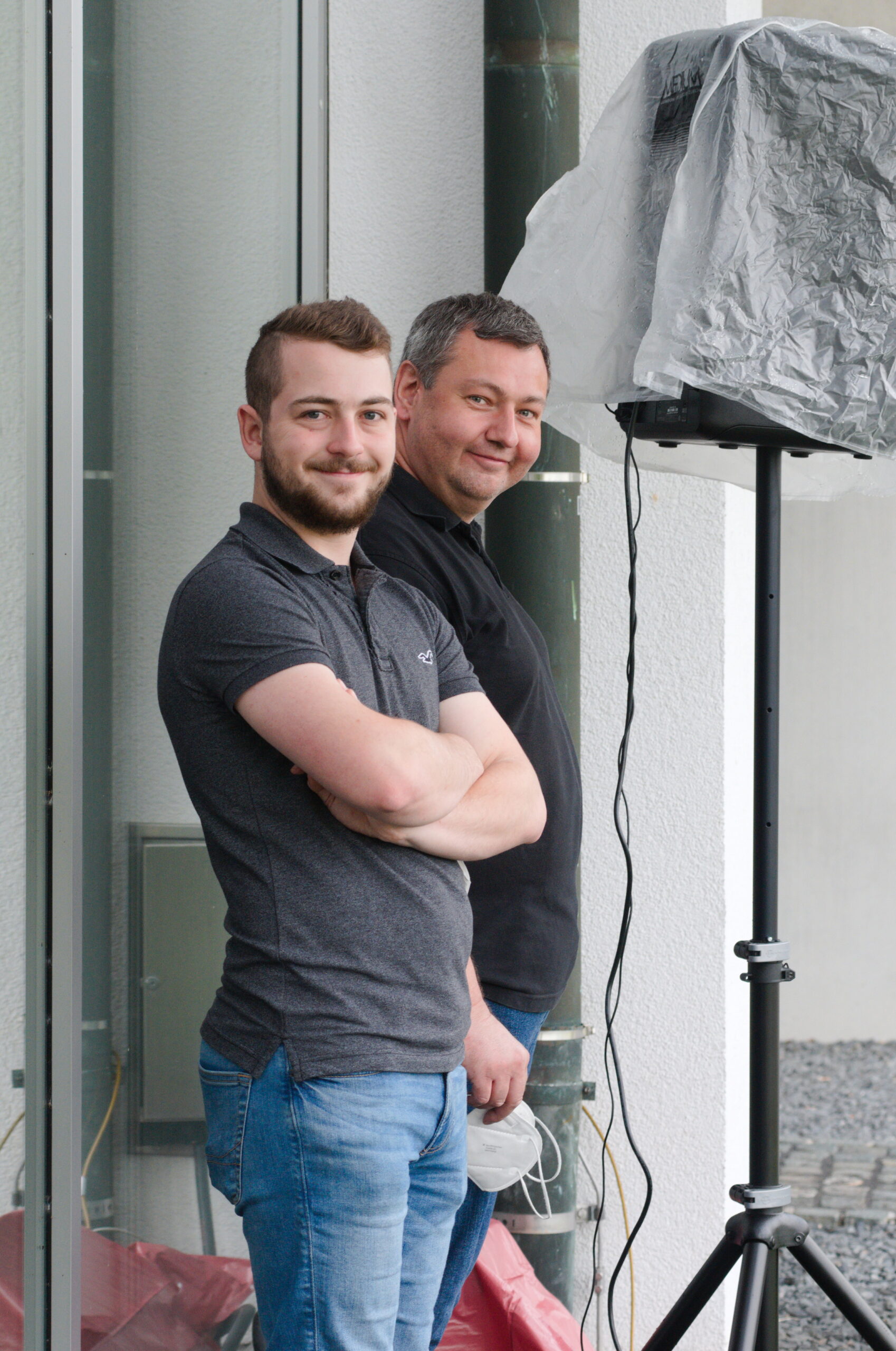 Zwei Männer stehen vor einem Gebäude und lächeln. Der bärtige Mann links trägt ein graues Hemd und Jeans, während sein Begleiter ein schwarzes Hemd trägt. Sie stehen neben einem abgedeckten elektronischen Gerät auf einem Stativ in der Nähe der Hochschule Coburg.