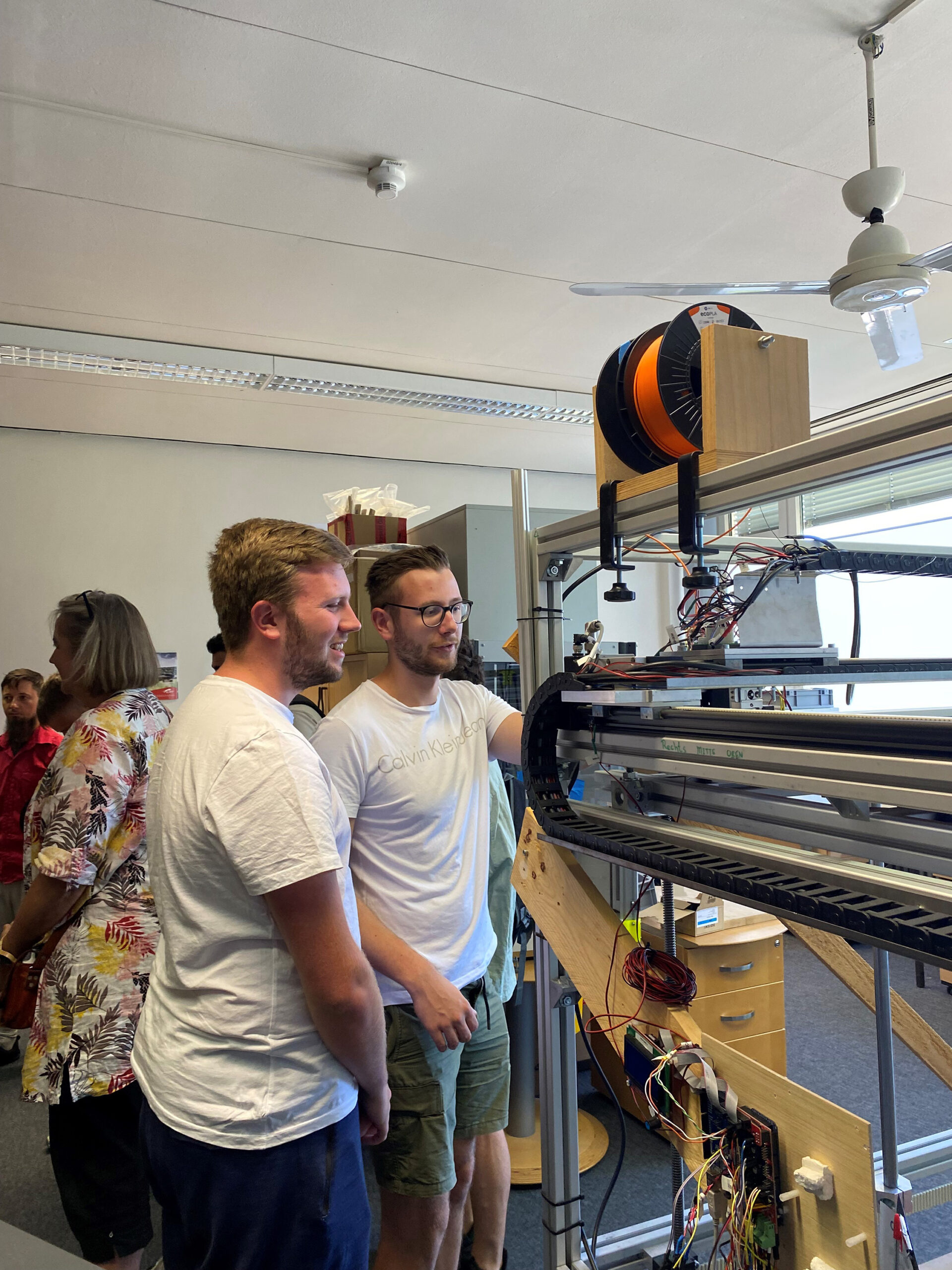 Zwei Menschen in weißen Hemden bedienen in einem Raum der Hochschule Coburg einen großen 3D-Drucker, im Hintergrund weitere. Die mit Spulen und diverser Elektronik ausgestattete Maschine schafft eine Atmosphäre, die zugleich locker und lehrreich ist.