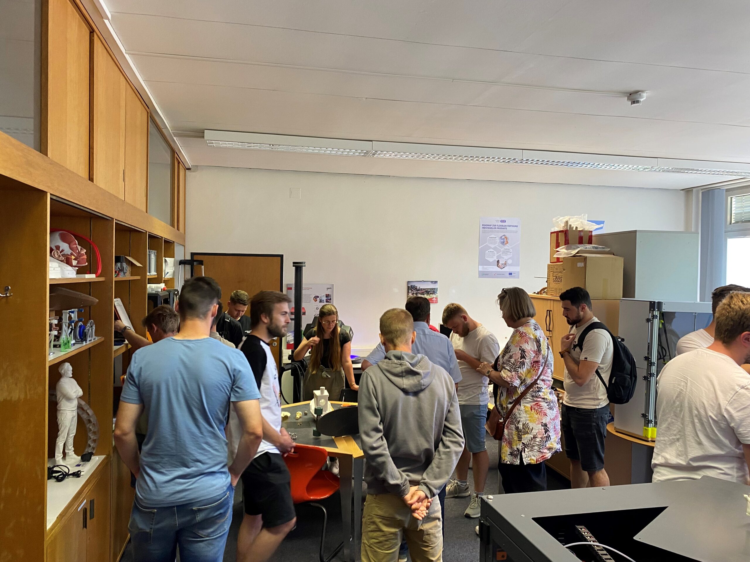 Eine Gruppe von Menschen versammelt sich in einem Raum der Hochschule Coburg, umgeben von Regalen und verschiedenen Gegenständen. Einige Personen konzentrieren sich auf einen 3D-Drucker im Vordergrund. Die Atmosphäre ist locker und doch engagiert, Poster schmücken die Wände.