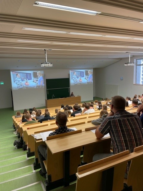 Ein Hörsaal der Hochschule Coburg ist voller Studenten. Sie sitzen an Holztischen und blicken nach vorne, wo auf einem großen Bildschirm eine Präsentation läuft. Der Raum mit seinem grünen Boden ist durch Deckenlampen gut beleuchtet, während mehrere Studenten fleißig Notizen machen.