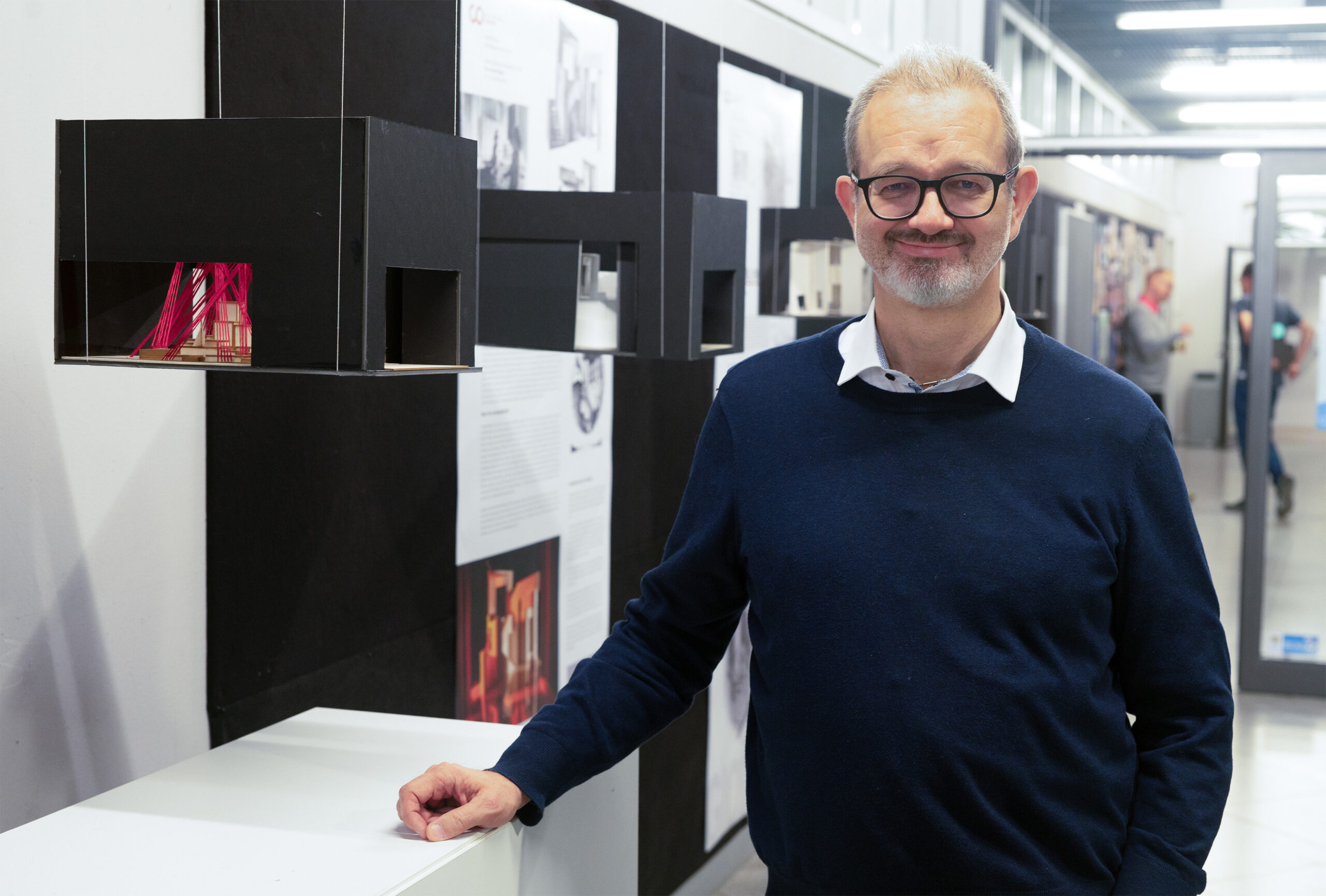 Ein Mann mit Brille und Bart, der den Stolz der Hochschule Coburg zur Schau stellt, lächelt, während er in einem Ausstellungsraum voller Architekturmodelle und Schautafeln steht. Er trägt einen marineblauen Pullover über einem weißen Hemd und verkörpert damit die Essenz akademischer Exzellenz.