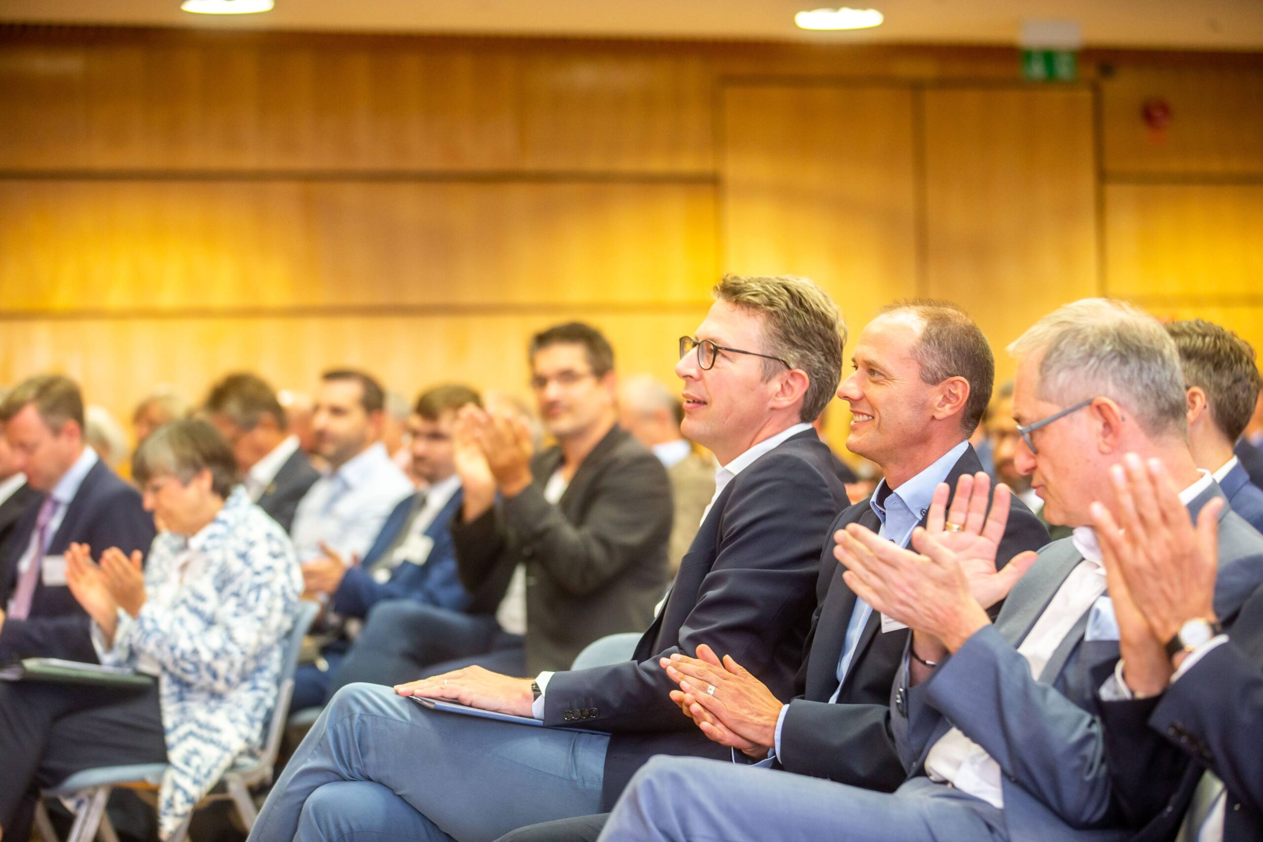 In einem Konferenzraum der Hochschule Coburg klatscht eine Gruppe von Menschen in Business-Kleidung, einige zeigen aufmerksame Mienen. Im Vordergrund stehen zwei Männer in Anzügen, im Hintergrund sieht man Reihen engagierter Teilnehmer.