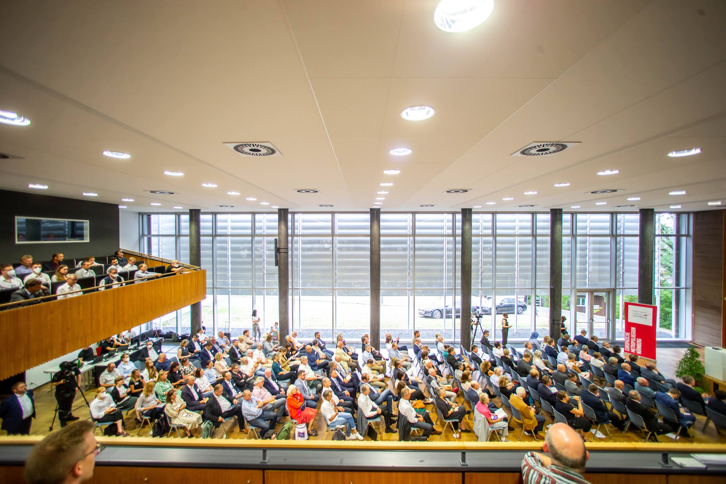 Weitwinkelansicht eines Hörsaals an der Hochschule Coburg, in dem Menschen in Reihen sitzen und aufmerksam einer Präsentation zuschauen. Der Raum verfügt über große Fenster, Holzvertäfelungen und Sitzreihen.