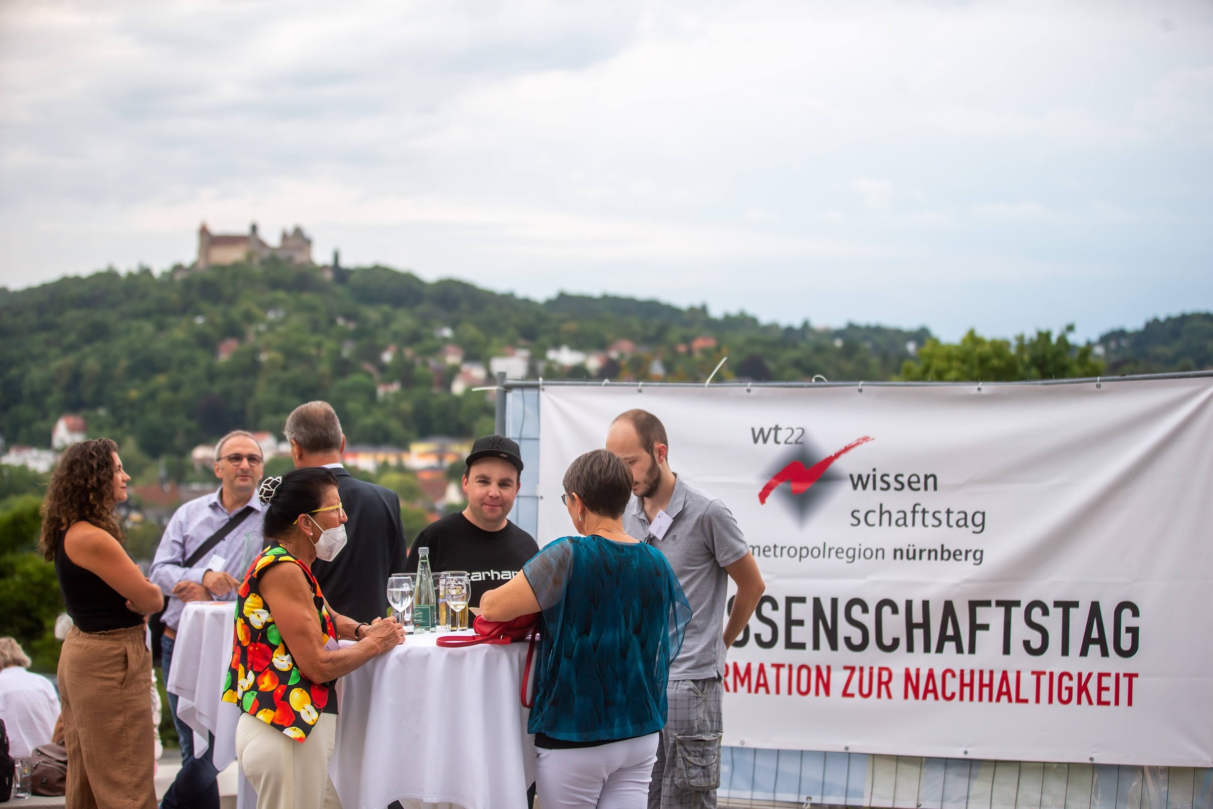 Eine Gruppe von Mitarbeitern der Hochschule Coburg sitzt bei einer Veranstaltung im Freien an Stehtischen. Ein Banner in deutscher Sprache unterstreicht das Thema Nachhaltigkeit. Sie genießen den malerischen Blick auf eine Hügellandschaft mit einer Burg in der Ferne im Hintergrund.