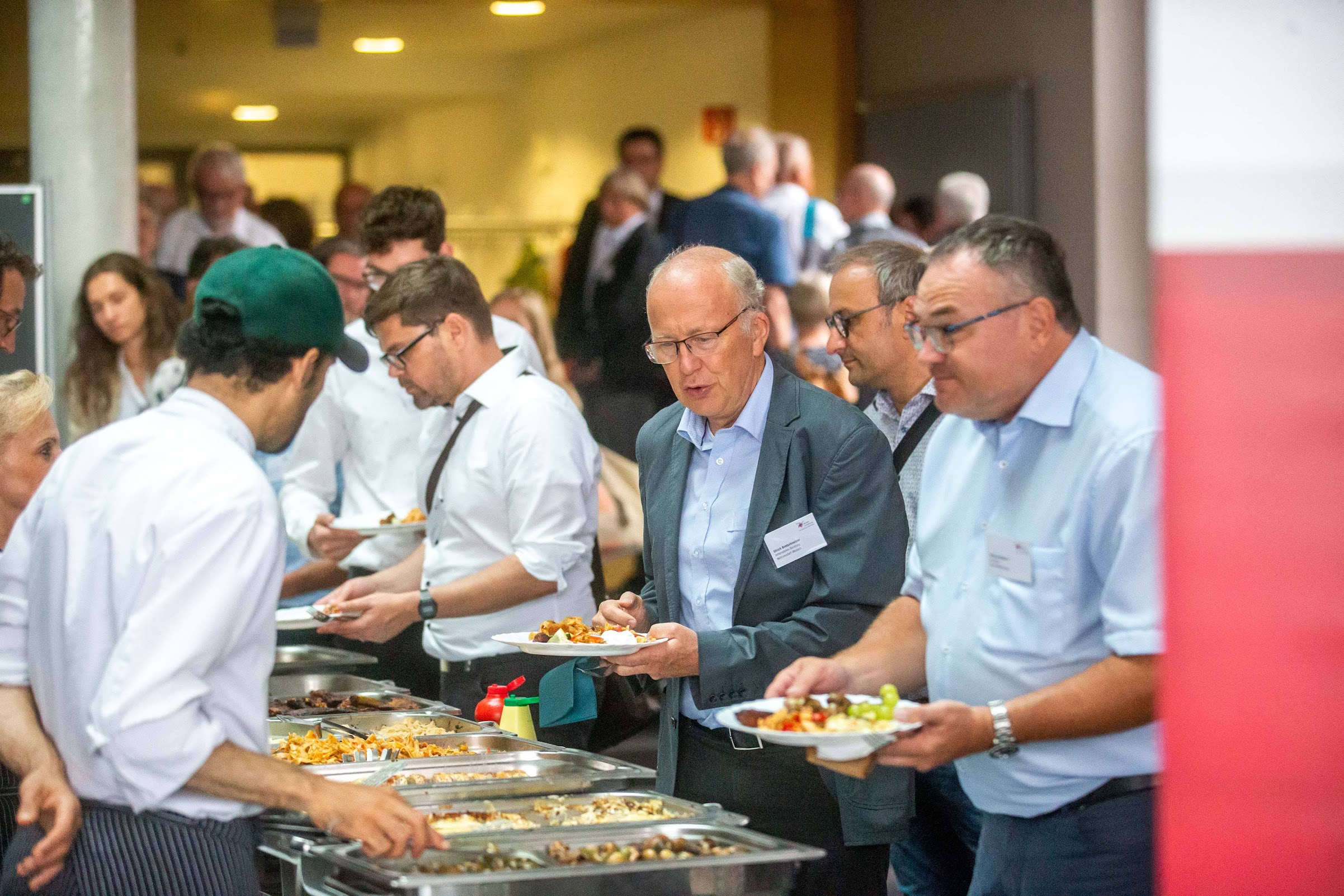 Eine Gruppe von Menschen steht an einem Buffettisch Schlange und bedient sich selbst. Mehrere Männer tragen hellblaue Hemden und tragen Namensschilder der Hochschule Coburg. Ein Koch in weißer Uniform und grüner Mütze hilft ihnen. Es scheint sich um eine Catering-Veranstaltung zu handeln.