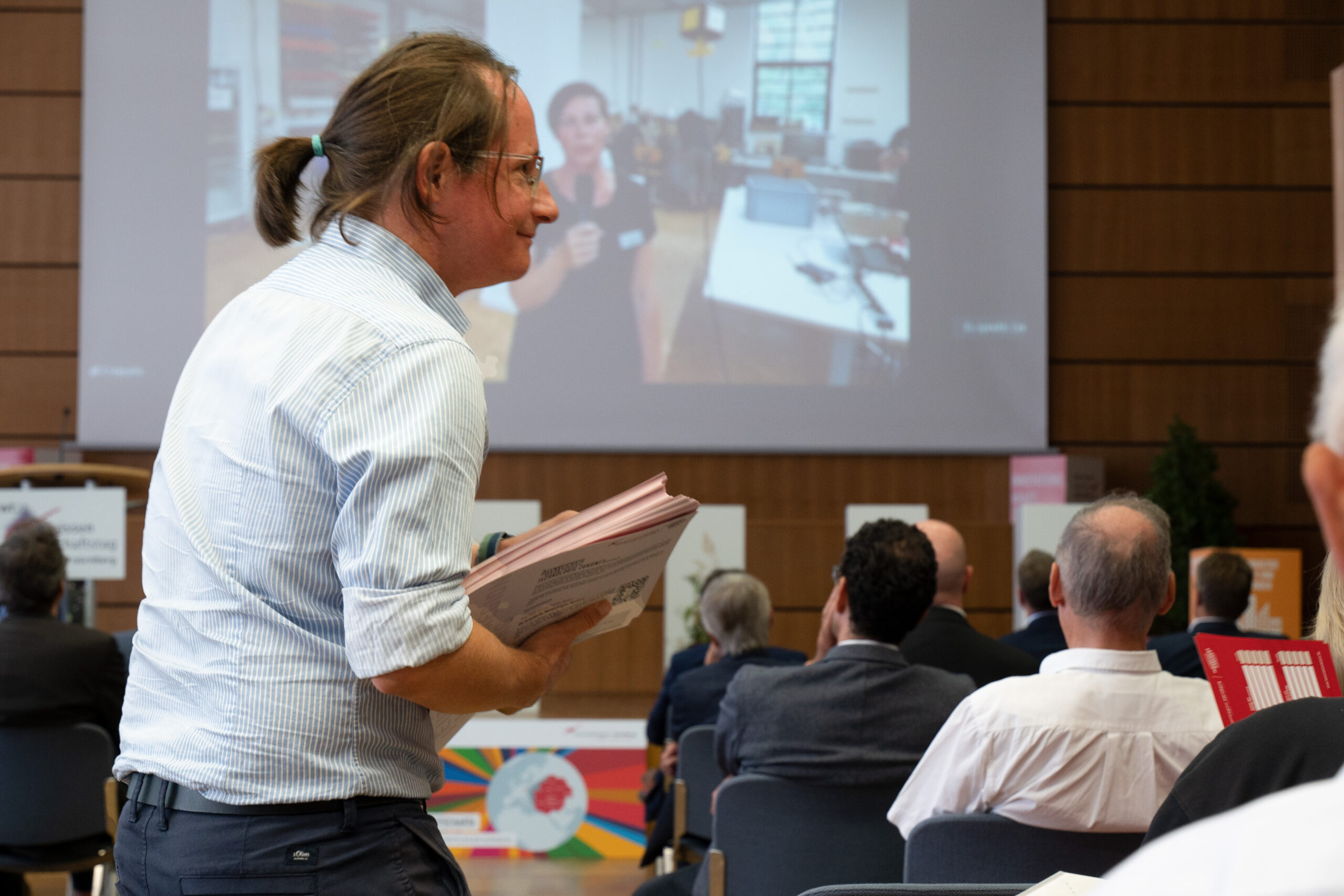 Eine Person mit zurückgebundenem langem Haar steht und hält Papiere während einer Präsentation an der Hochschule Coburg. Im Hintergrund sitzen Menschen vor einem großen Bildschirm, auf dem eine Frau spricht. Der Raum ist mit Holzvertäfelungen und verschiedenen Postern ausgestattet.