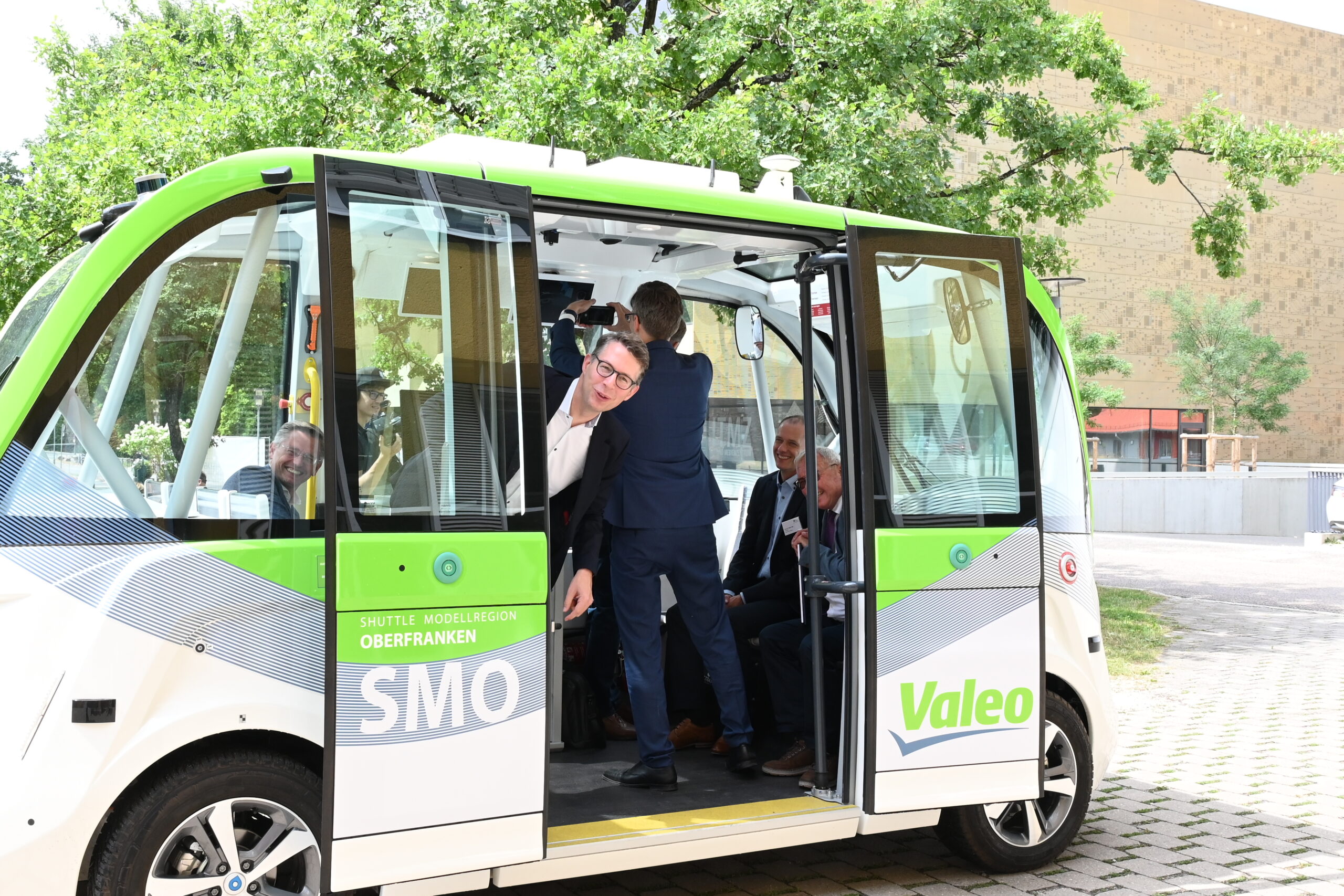 Ein Mann lehnt sich aus der offenen Tür eines kleinen grün-weißen autonomen Shuttles mit den Logos „SMO“ und „Valeo“, Teil des innovativen Transportsystems der Hochschule Coburg. Im Inneren sitzen einige Leute. Das Fahrzeug steht auf einer gepflasterten Fläche, umgeben von Grünflächen und Gebäuden.