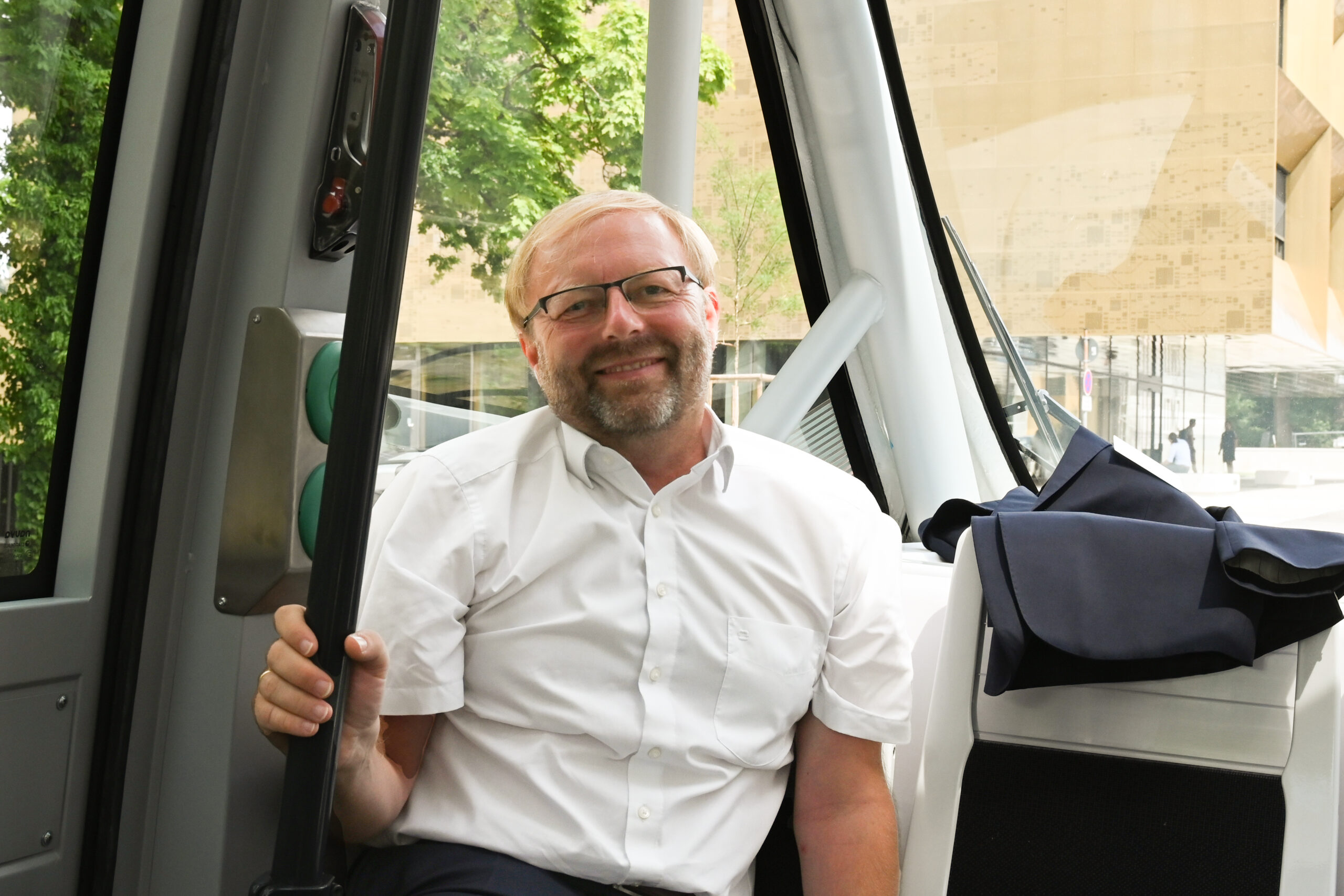 Ein Mann mit Brille, Bart und weißem Hemd sitzt lächelnd in einem modernen Fahrzeug. Helles Tageslicht erhellt die Szene und gibt durch das Fenster den Blick auf die Hochschule Coburg und üppiges Grün frei.