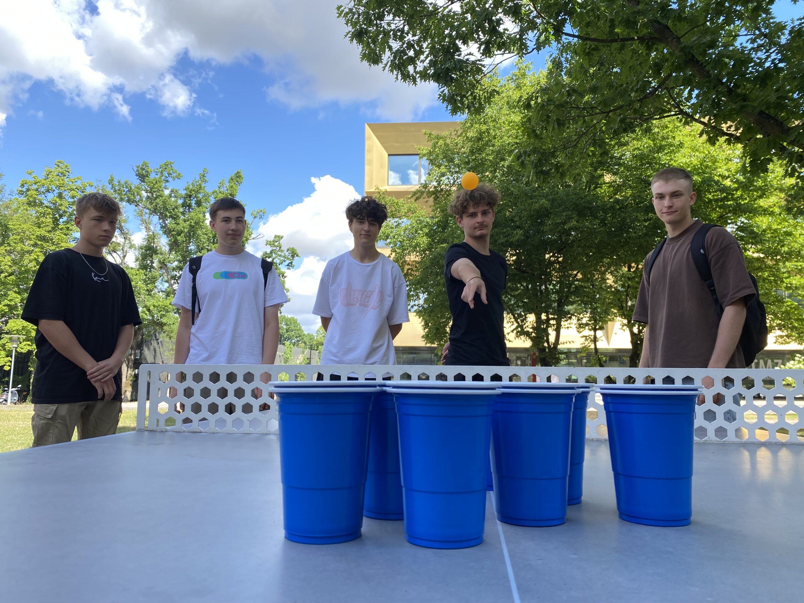 Fünf junge Männer stehen im Freien an der Hochschule Coburg um einen Tisch und spielen ein Spiel mit blauen Plastikbechern und einem Tischtennisball. Einer von ihnen versucht, den Ball in die Becher zu werfen. Im Hintergrund sind Bäume und ein Gebäude zu sehen, die zur lebhaften Campusatmosphäre beitragen.