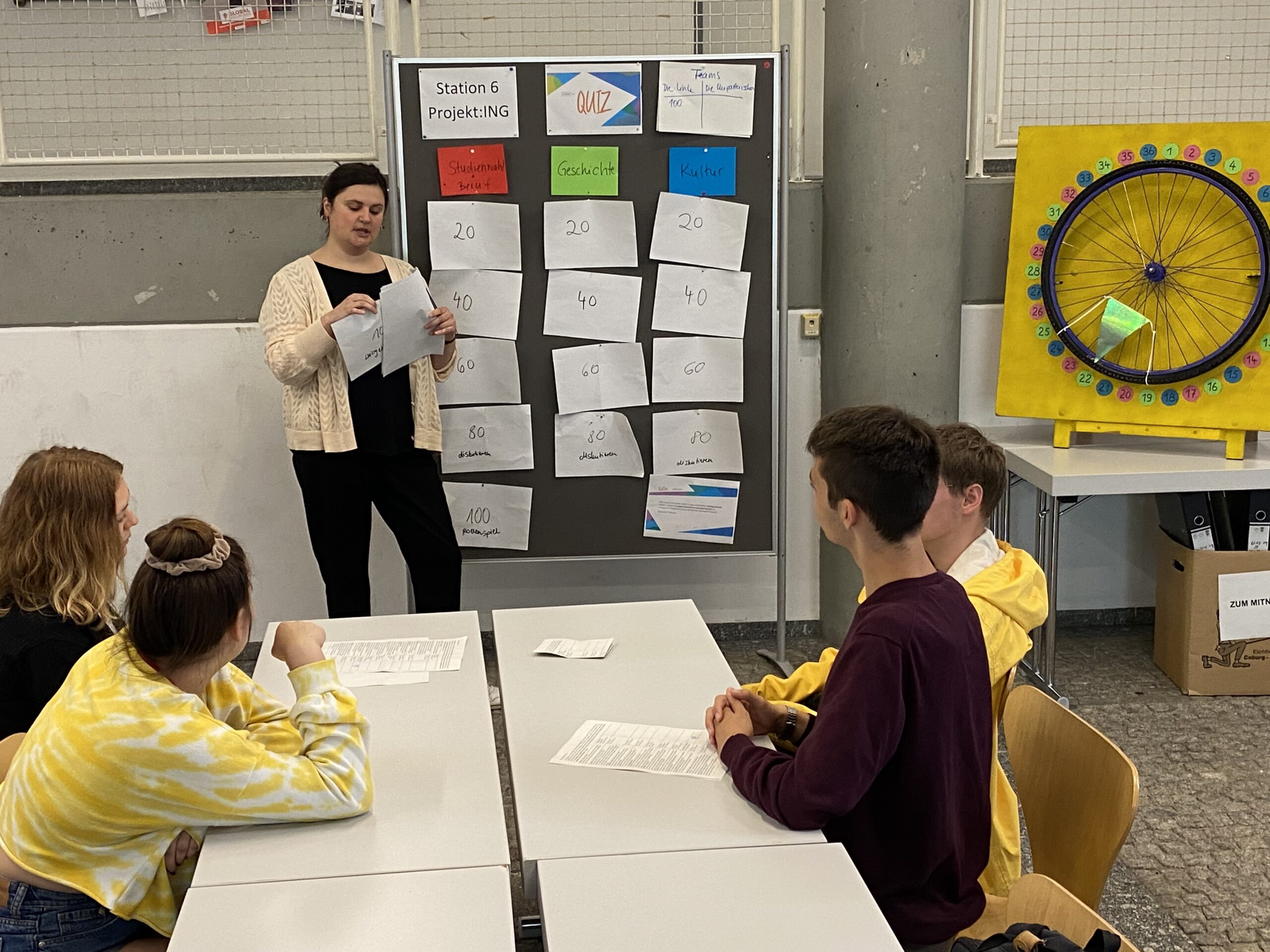 Eine Frau steht vor einer Gruppe sitzender Studenten an der Hochschule Coburg und zeigt auf eine Quiztafel mit nummerierten Karten. Die Kulisse scheint ein Klassenzimmer voller Tische und Stühle zu sein, während sich im Hintergrund ein buntes Rad lebhaft dreht.