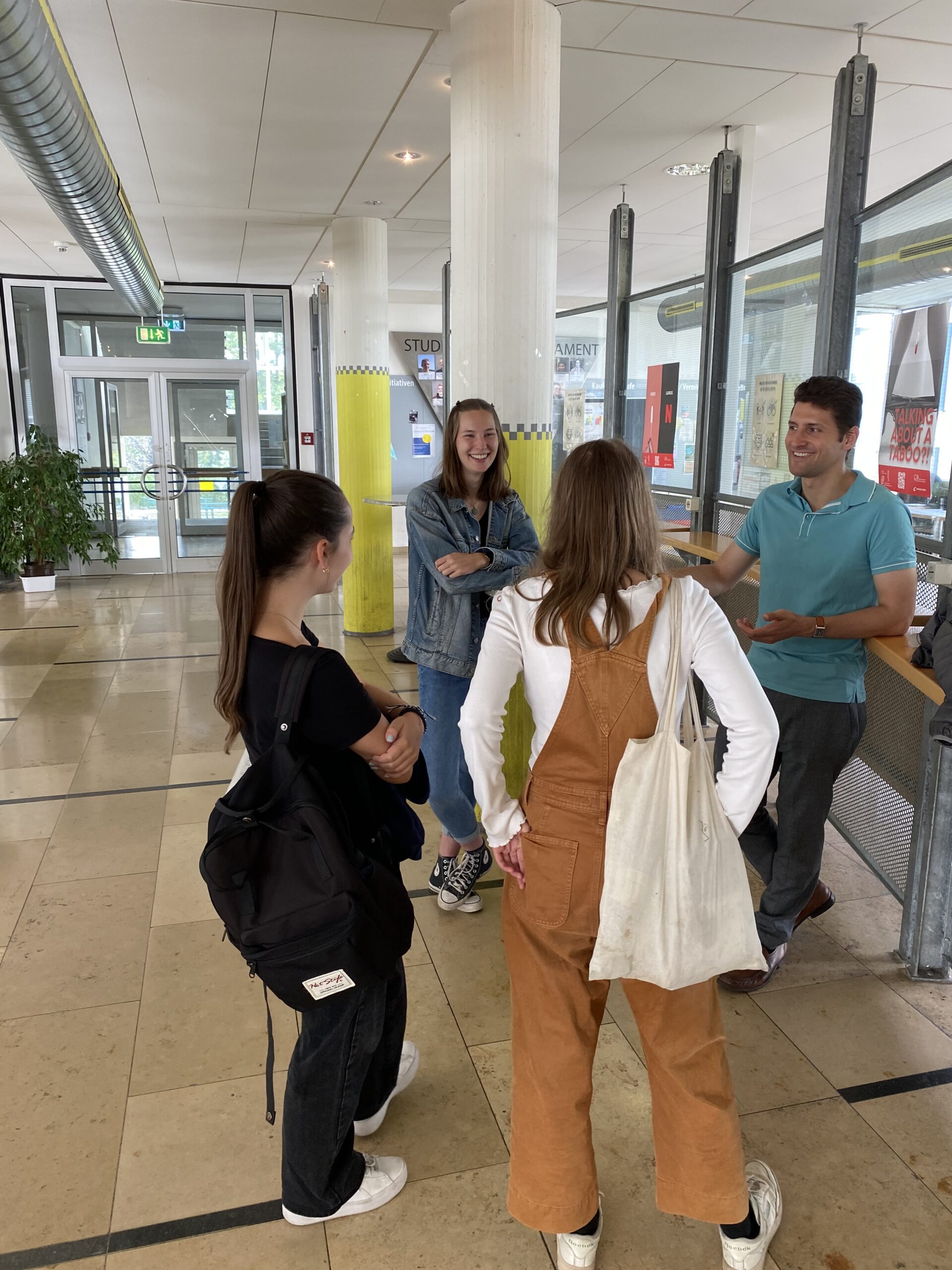 Vier Menschen stehen in einem Flur der Hochschule Coburg und unterhalten sich. Drei sind junge Frauen, einer ist ein junger Mann. Sie sind leger gekleidet, mit Rucksäcken und Taschen. Der Flur ist gut beleuchtet, mit großen Fenstern und moderner Einrichtung.