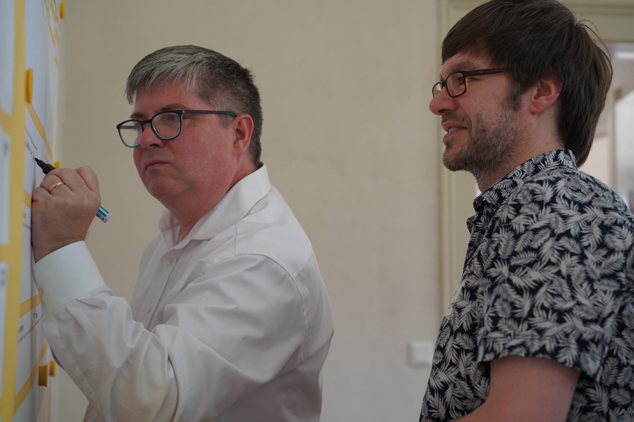 Zwei Männer stehen an einem Whiteboard in der Hochschule Coburg. Der Mann links, mit Brille und weißem Hemd, schreibt mit einem Marker. Der Mann rechts, ebenfalls mit Brille, trägt ein gemustertes Hemd und beobachtet aufmerksam die Innenumgebung.