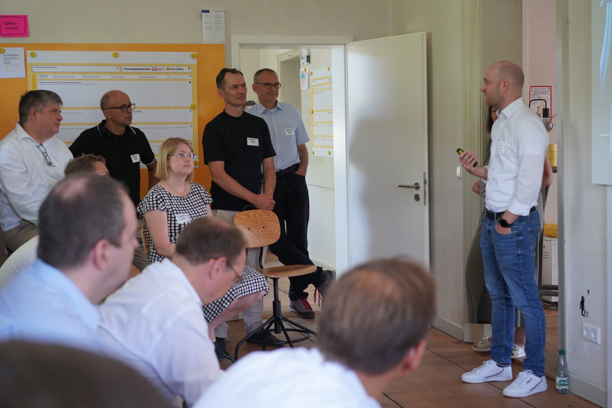 Ein Mann steht vor einer Gruppe und hält einen Vortrag an der Hochschule Coburg. Das Publikum, sitzend und stehend, hört aufmerksam zu. An den Wänden sind ein Whiteboard und Diagramme zu sehen. Die Kulisse scheint ein Büro oder ein Besprechungsraum zu sein.