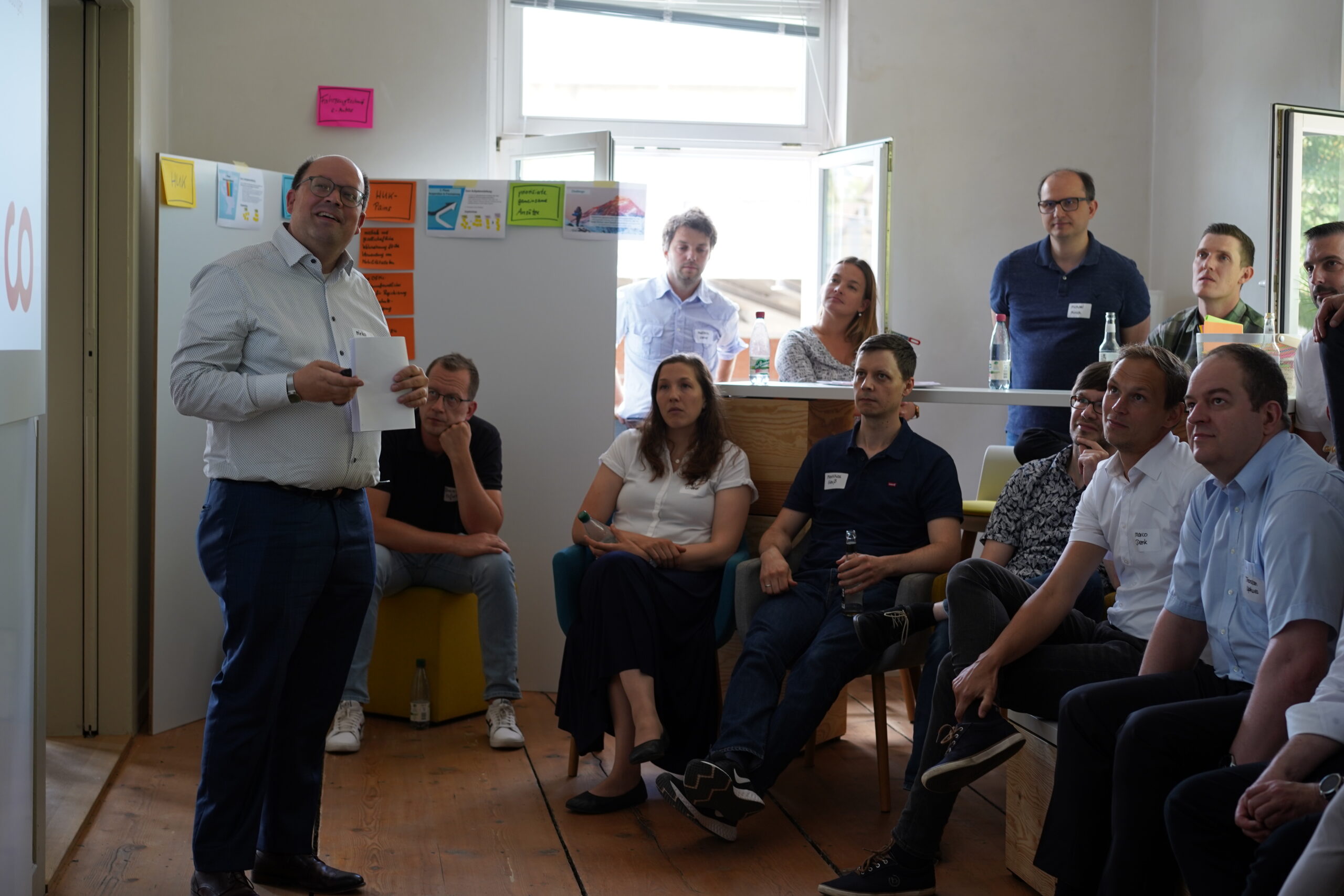 In dem gut beleuchteten Raum der Hochschule Coburg hört eine Gruppe von Menschen einem Redner aufmerksam zu, der ein Referat in der Hand hält. Bunte Haftnotizen zieren die Whiteboards, während natürliches Licht durch die offenen Fenster strömt. Einige sitzen, andere stehen, aber alle wirken interessiert.