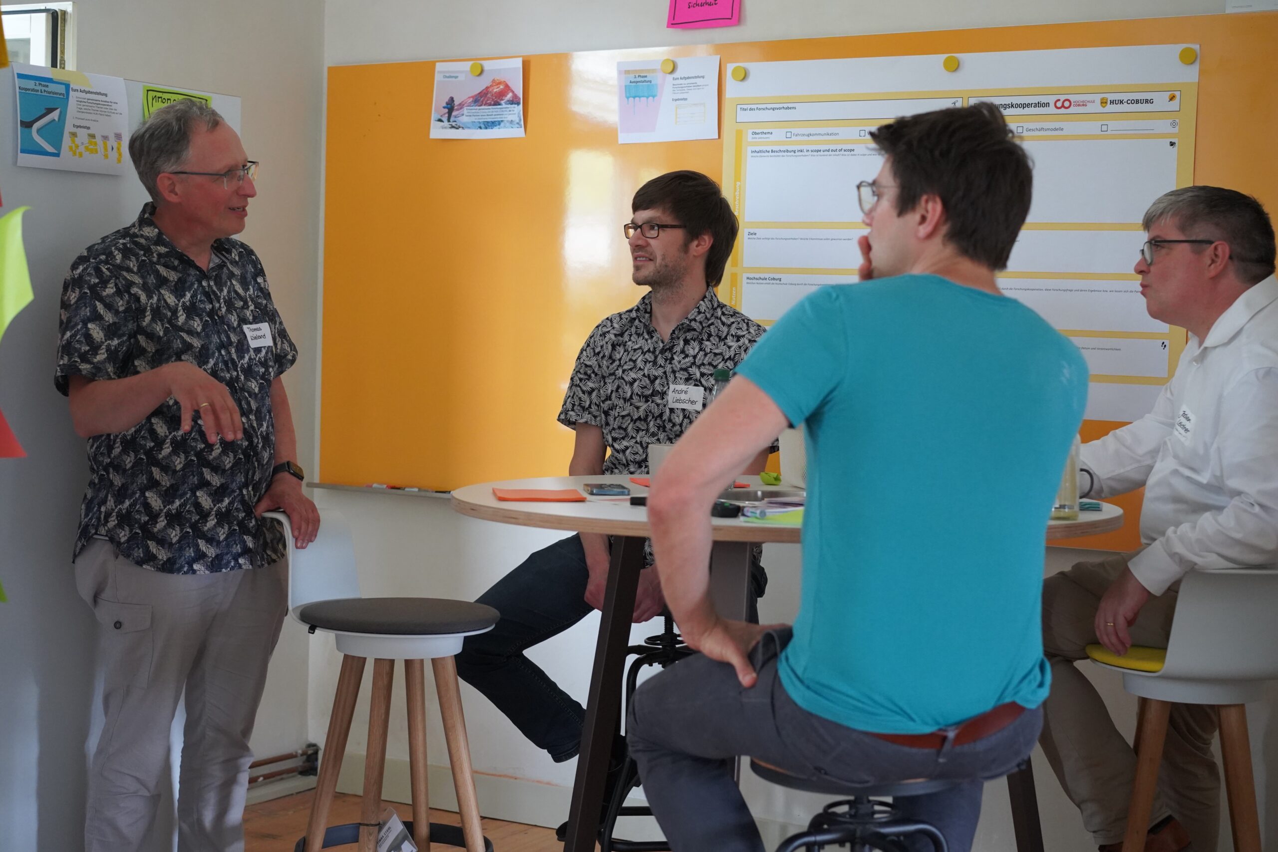 Vier Männer diskutieren in einem hellen Raum der Hochschule Coburg. Ein Mann spricht im Stehen, die anderen sitzen um einen hohen Tisch herum, an dessen Wänden Büroplakate hängen, und scheinen sich in einer gemeinsamen Besprechung zu befinden.