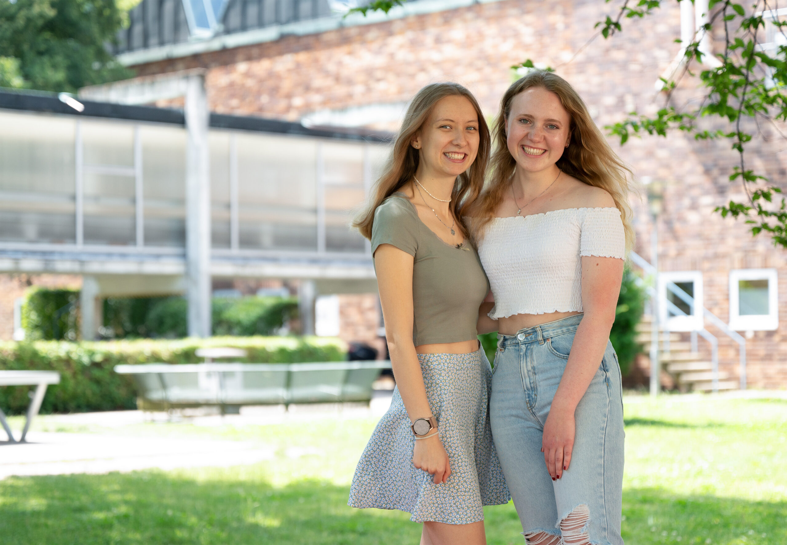 Two young women stand outdoors, smiling at the camera in front of Hochschule Coburg. They are dressed casually in summer outfits, with greenery and a modern building in the background on a sunny day.