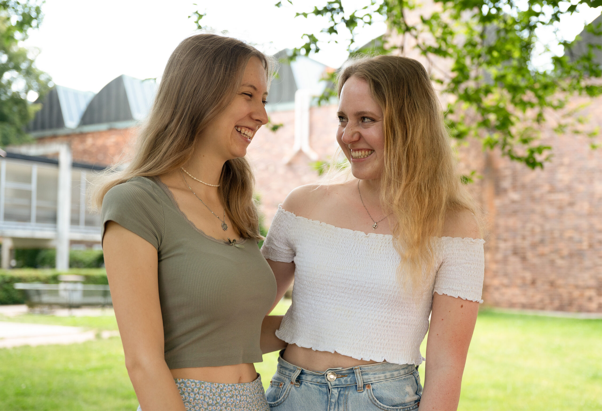 In front of a brick building at Hochschule Coburg, two women smile warmly at each other. One sports a green top with patterned pants, while the other is in a white off-shoulder top and denim shorts, surrounded by lush greenery.