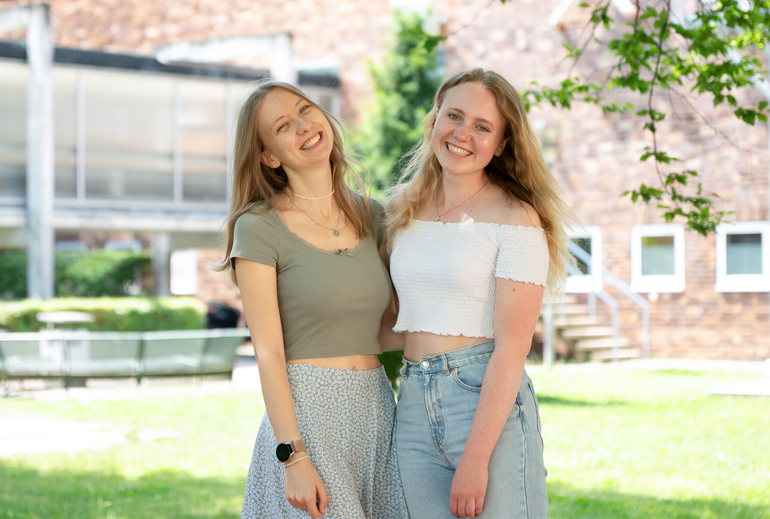 Two young women, one in a green top and floral skirt and the other in a white off-shoulder top and jeans, smile brightly under the sun, standing amidst greenery before a brick building at Hochschule Coburg.