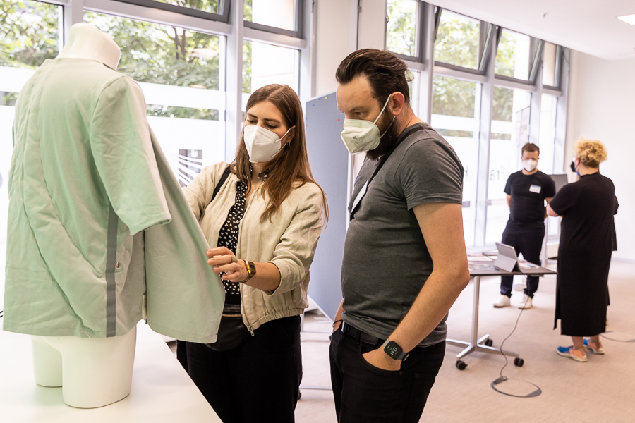 Zwei maskierte Personen untersuchen ein Kleidungsstück an einer Schaufensterpuppe in einem modernen Raum mit großen Fenstern an der Hochschule Coburg. Im Hintergrund unterhalten sich zwei weitere Personen mit Masken an einem Tisch. Tische mit Laptops und Papieren sind zu sehen und spiegeln die dynamische Bildungsumgebung wider.