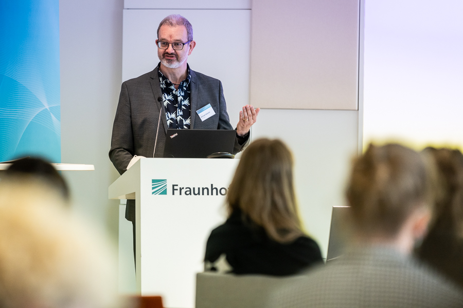 Ein Mann spricht während einer Präsentation auf einem Podium. Er trägt einen dunklen Anzug und ein gemustertes Hemd. Das Fraunhofer-Emblem ist deutlich zu sehen. Das unscharfe Publikum lässt vermuten, dass Teilnehmer möglicherweise mit der Hochschule Coburg in Verbindung stehen.