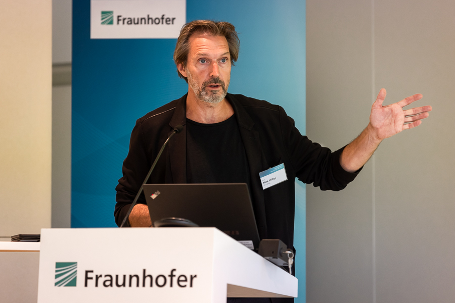 Ein Mann mit Bart und schwarzer Kleidung spricht an einem Rednerpult mit der Aufschrift „Fraunhofer“ und gestikuliert mit seiner rechten Hand. Ein blauer Hintergrund, auf dem sowohl das Fraunhofer-Logo als auch die Zugehörigkeit zur Hochschule Coburg zu sehen sind, umrahmt ihn. Ein Laptop und ein Mikrofon stehen auf dem Rednerpult und erleichtern seine Präsentation.