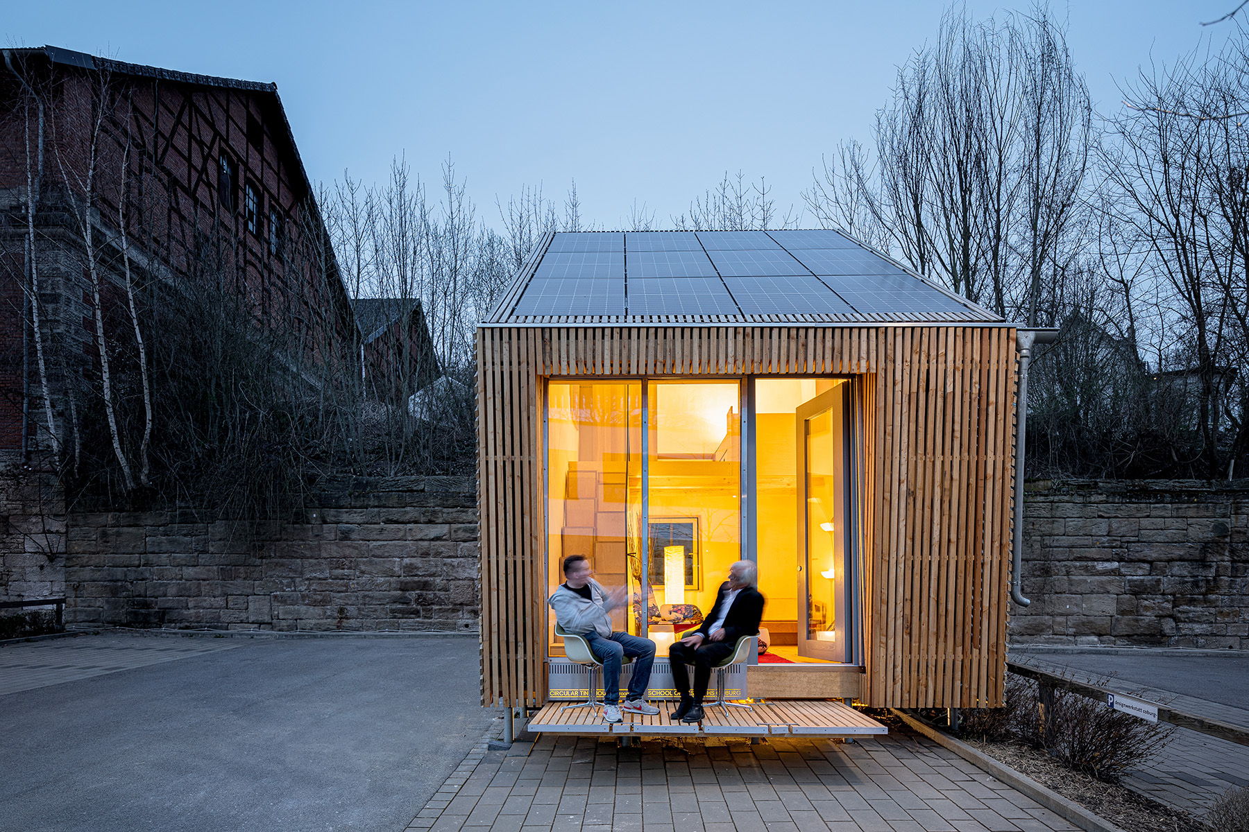 Ein kleines, modernes Holzhaus mit Solarmodulen auf dem Dach liegt in der Nähe der Hochschule Coburg. Zwei Menschen unterhalten sich auf der Veranda. Es ist Abend und das warme Licht im Inneren kontrastiert mit dem blauen Himmel. Im Hintergrund sind blattlose Bäume und ein Backsteingebäude zu sehen.