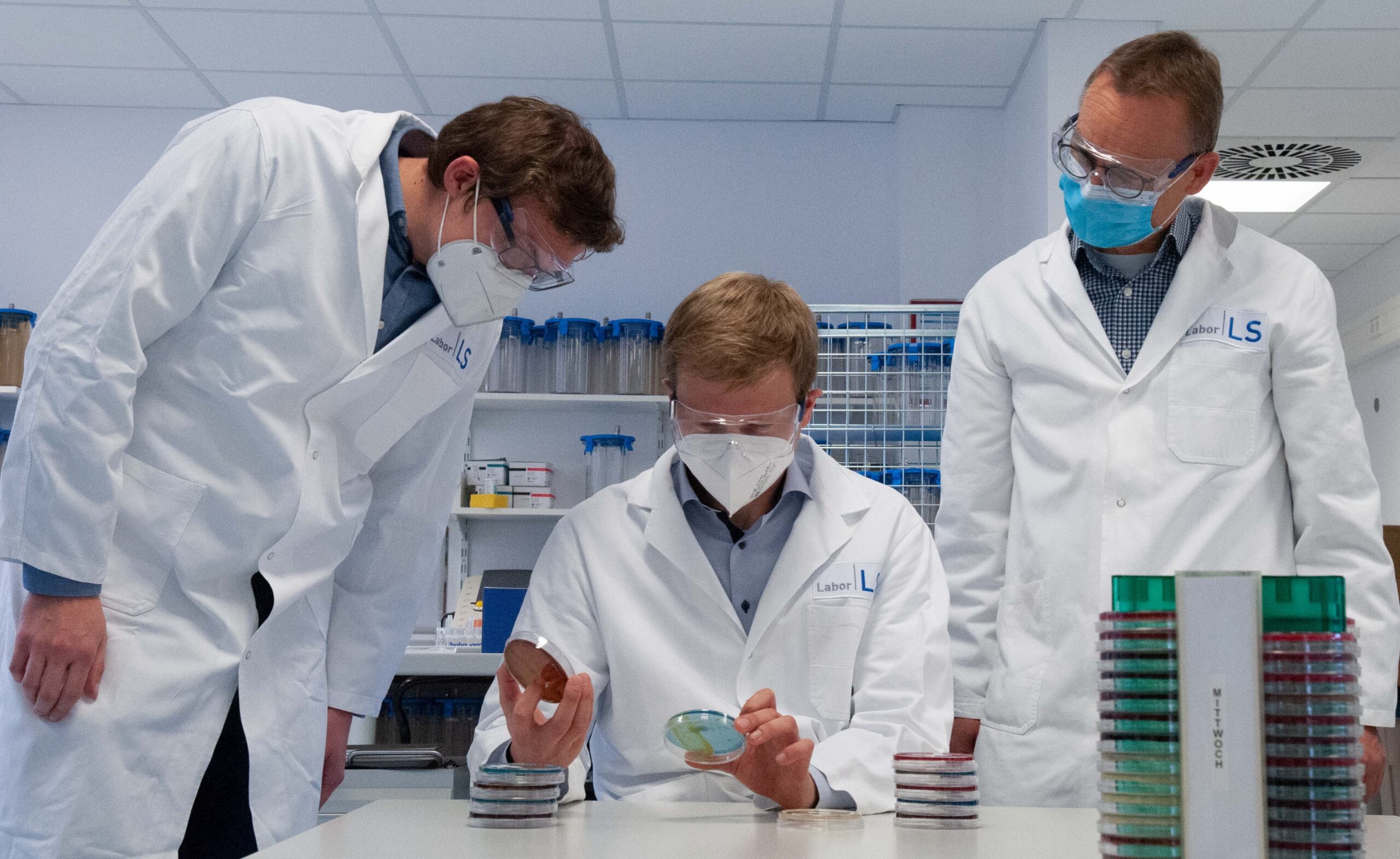 Drei Wissenschaftler in Laborkitteln und Masken untersuchen sorgfältig Petrischalen in einem Labor der Hochschule Coburg. Der Raum ist voller Regale und hochmoderner Geräte, auf dem Tisch liegen Stapel von Petrischalen.