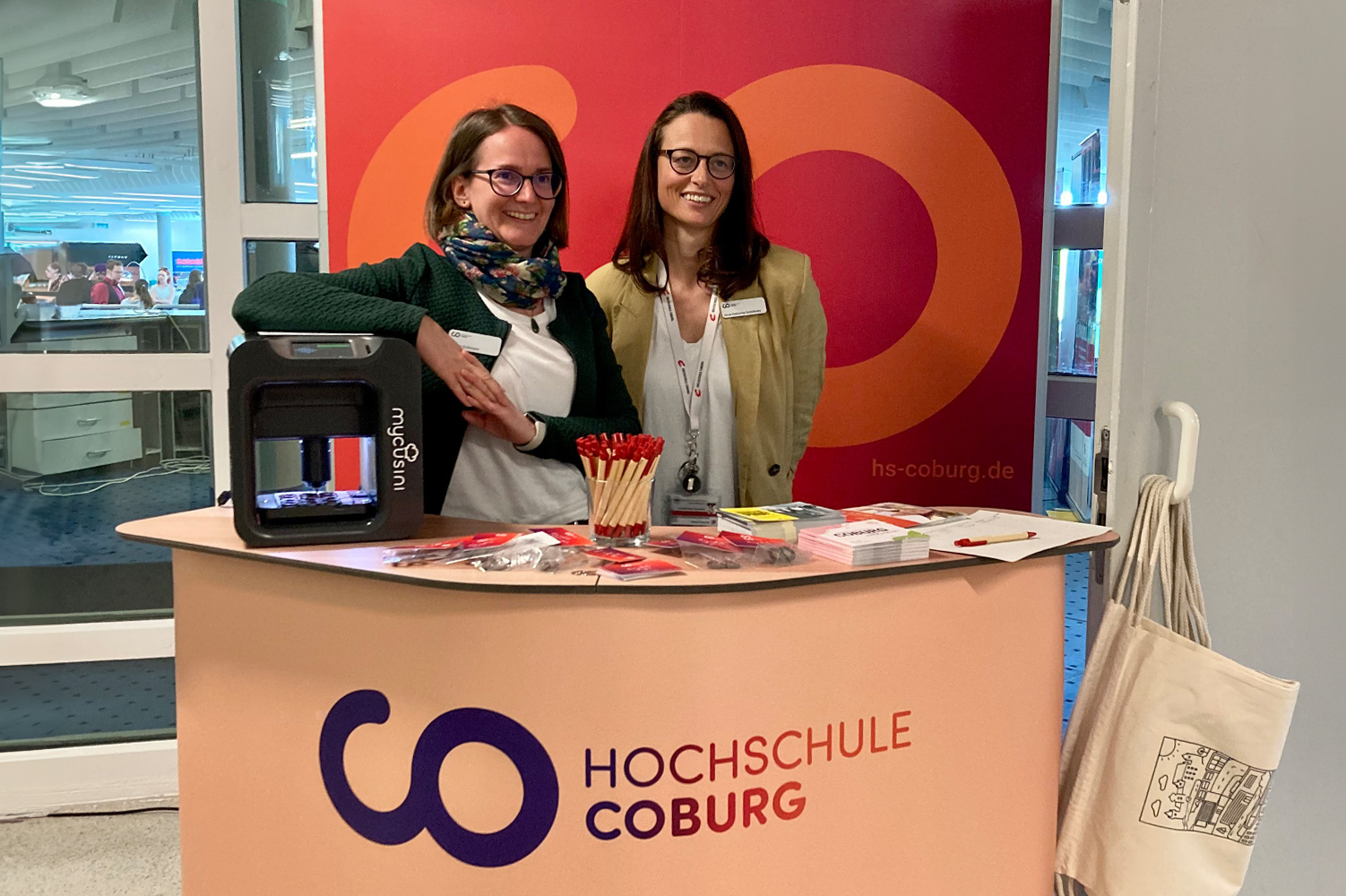 Two people stand behind an inviting Hochschule Coburg information booth, where brochures, pens, and a small 3D printer are on display. A tote bag hangs on the side. The backdrop proudly features Hochschule Coburg’s logo and website.