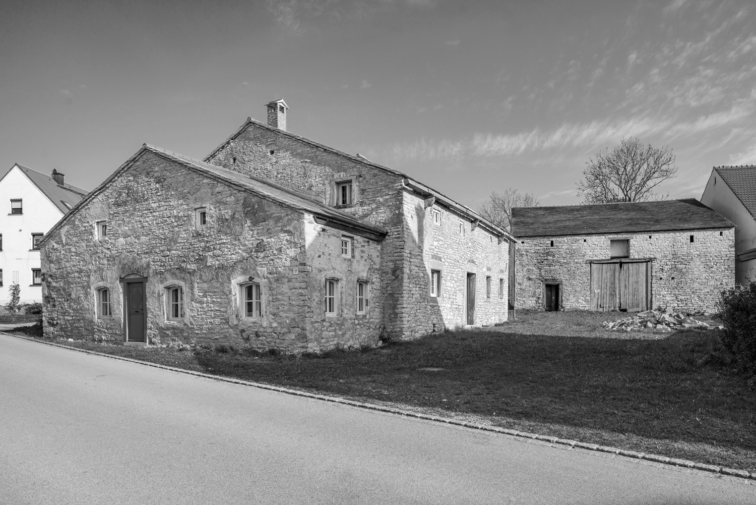 Ein Schwarzweißbild zeigt ein altes Bauernhaus aus Stein in der Nähe der Hochschule Coburg mit mehreren kleinen Fenstern und einer Holztür. Das Gebäude steht neben einer gepflasterten Straße, im Vordergrund von Gras eingerahmt und darüber von einem leicht bewölkten Himmel.