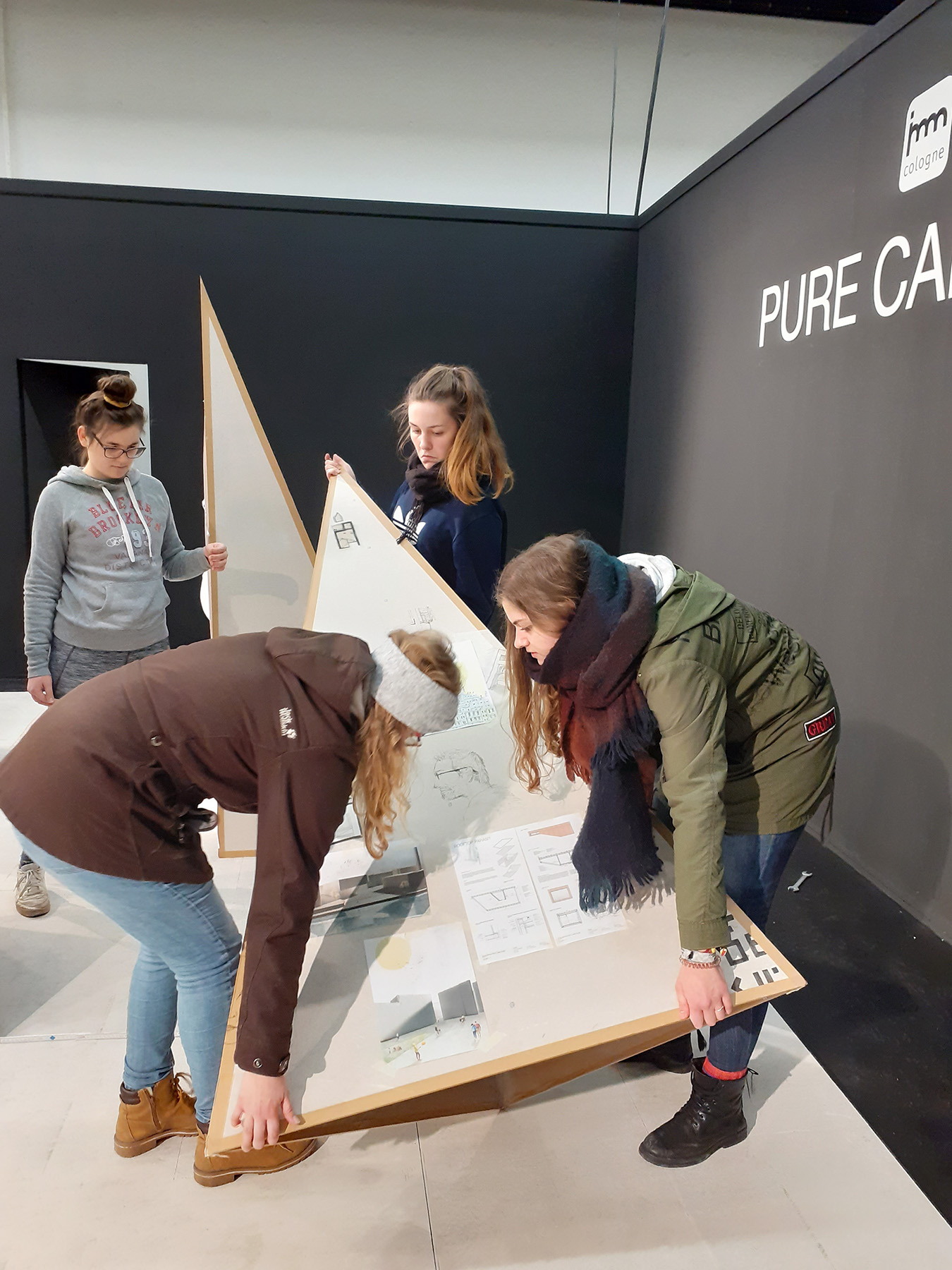 In einem Raum mit dunklen Wänden, in dem „PURE C“ teilweise sichtbar ist, bauen vier Personen eine große dreieckige Holzkonstruktion zusammen, möglicherweise für eine Ausstellung. Eine Person hält Anweisungen. Das minimalistische, moderne Design lässt Einflüsse der innovativen Architekturprinzipien der Hochschule Coburg erahnen.