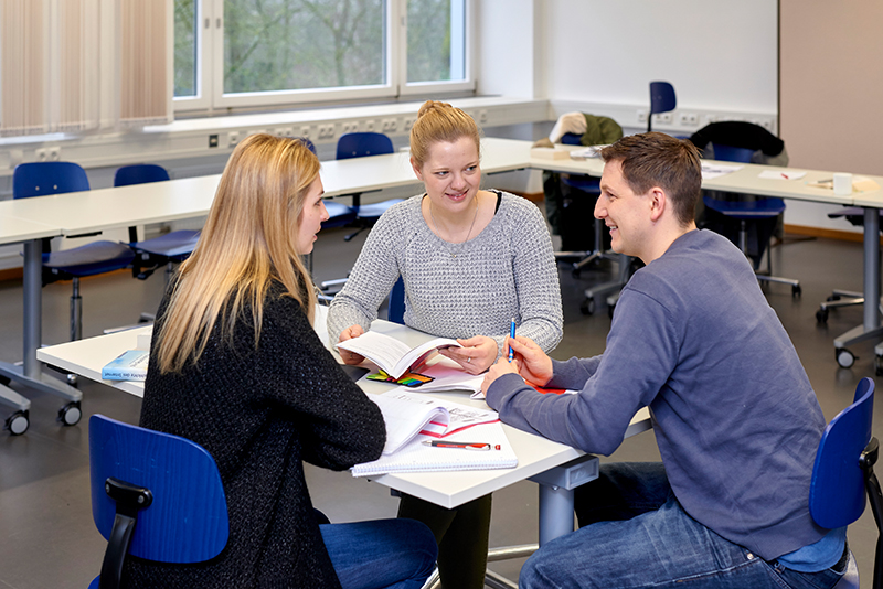 Drei Personen sitzen an einem Tisch im Hörsaal der Hochschule Coburg und diskutieren angeregt. Mit Notizbüchern und einem offenen Buch tauschen sie zwischen Tischen und Stühlen Ideen aus, im Hintergrund fällt Sonnenlicht durch große Fenster.