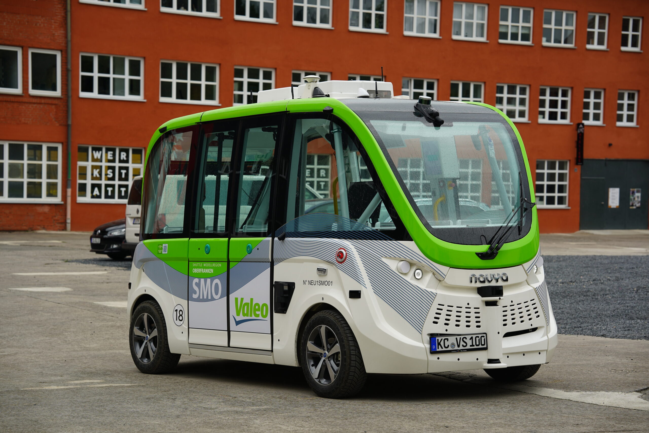 A small, driverless shuttle bus with a green and white design, boasting the Hochschule Coburg connection, is parked in an urban area. It features Valeo and Navya logos and sensors on the roof. A red brick building with multiple windows stands in the background.