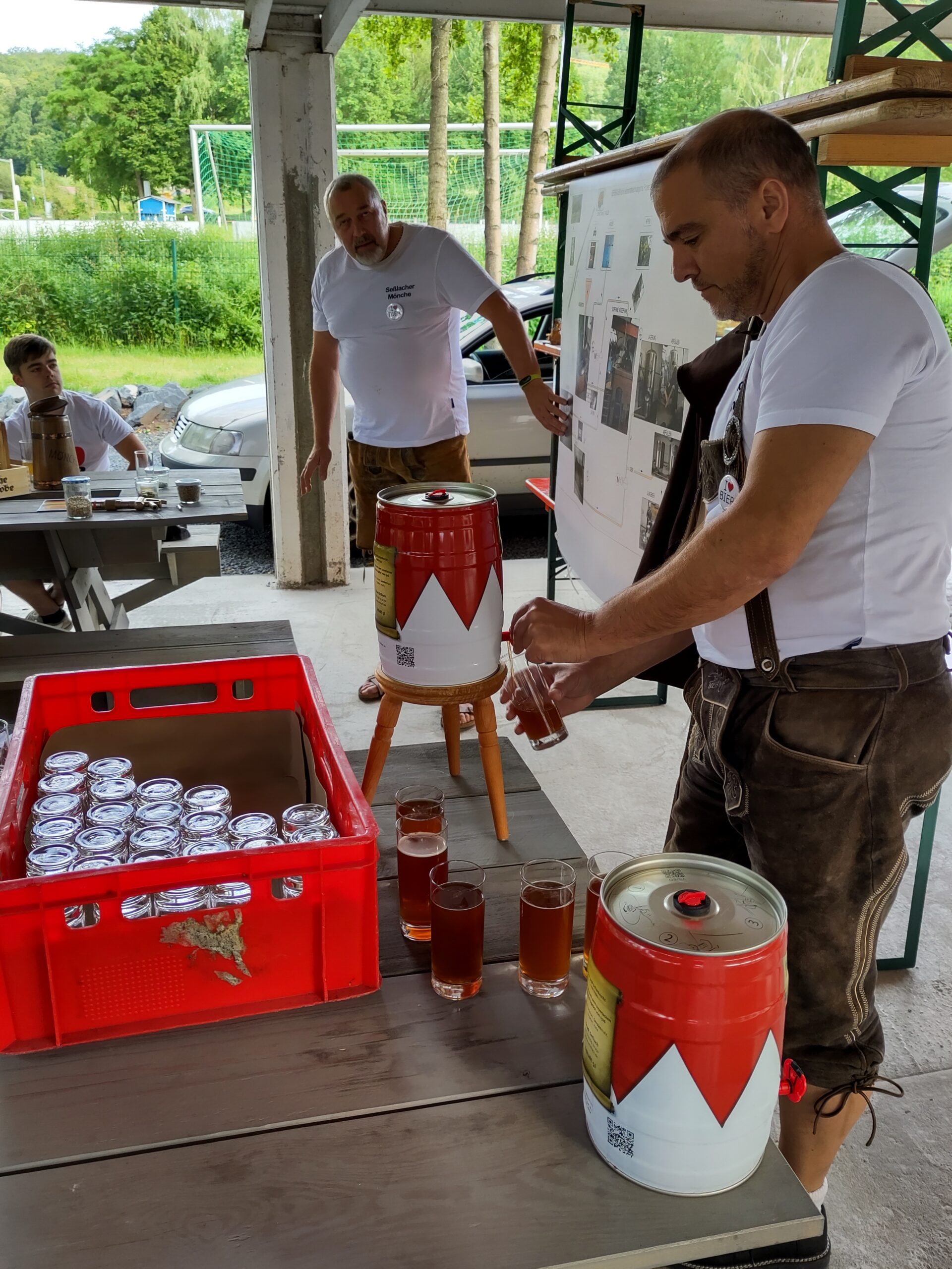 A man in lederhosen pours beer from a mini keg into glasses at an outdoor table adorned with a red crate of beer cans. Nearby, another man gestures toward a display board promoting Hochschule Coburg. In the background, a lush, green landscape completes the vibrant scene.