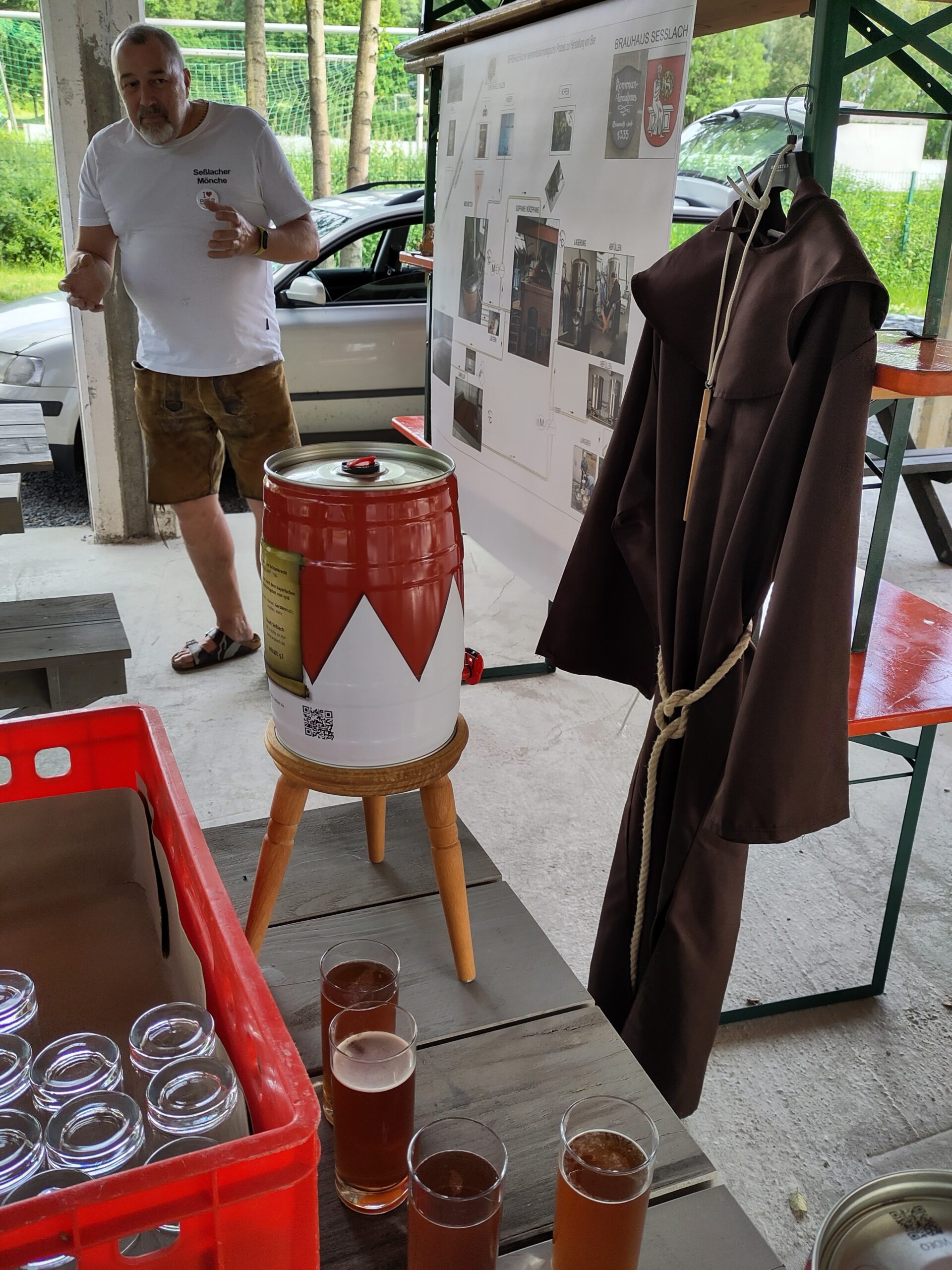 A man stands near a table with a barrel on a stool, surrounded by glasses of beer. A monk's robe hangs nearby. In the background, a poster showcases images and text, hinting at Hochschule Coburg's involvement in this casual outdoor event or tasting.