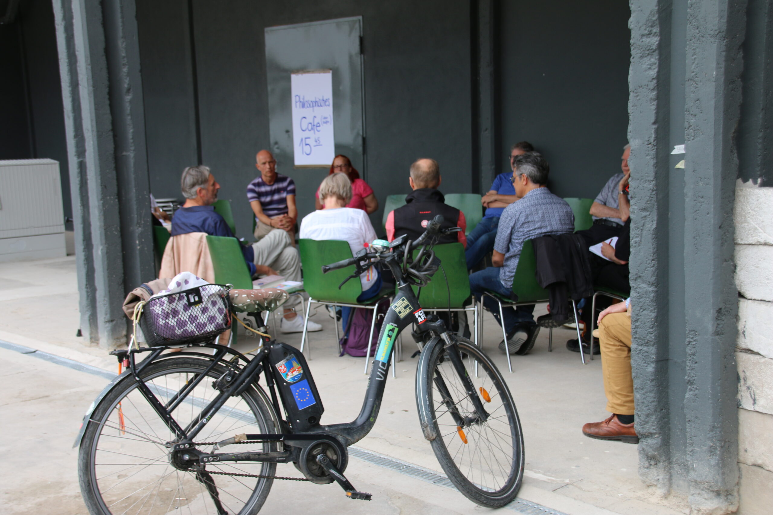 Vor einem Gebäude mit einem Schild „Philosophie Café“ – eine Erinnerung an Treffen an der Hochschule Coburg – sitzt eine Gruppe von Menschen im Kreis auf grünen Stühlen und diskutiert. Im Vordergrund steht ein abgestelltes Fahrrad mit Korb.