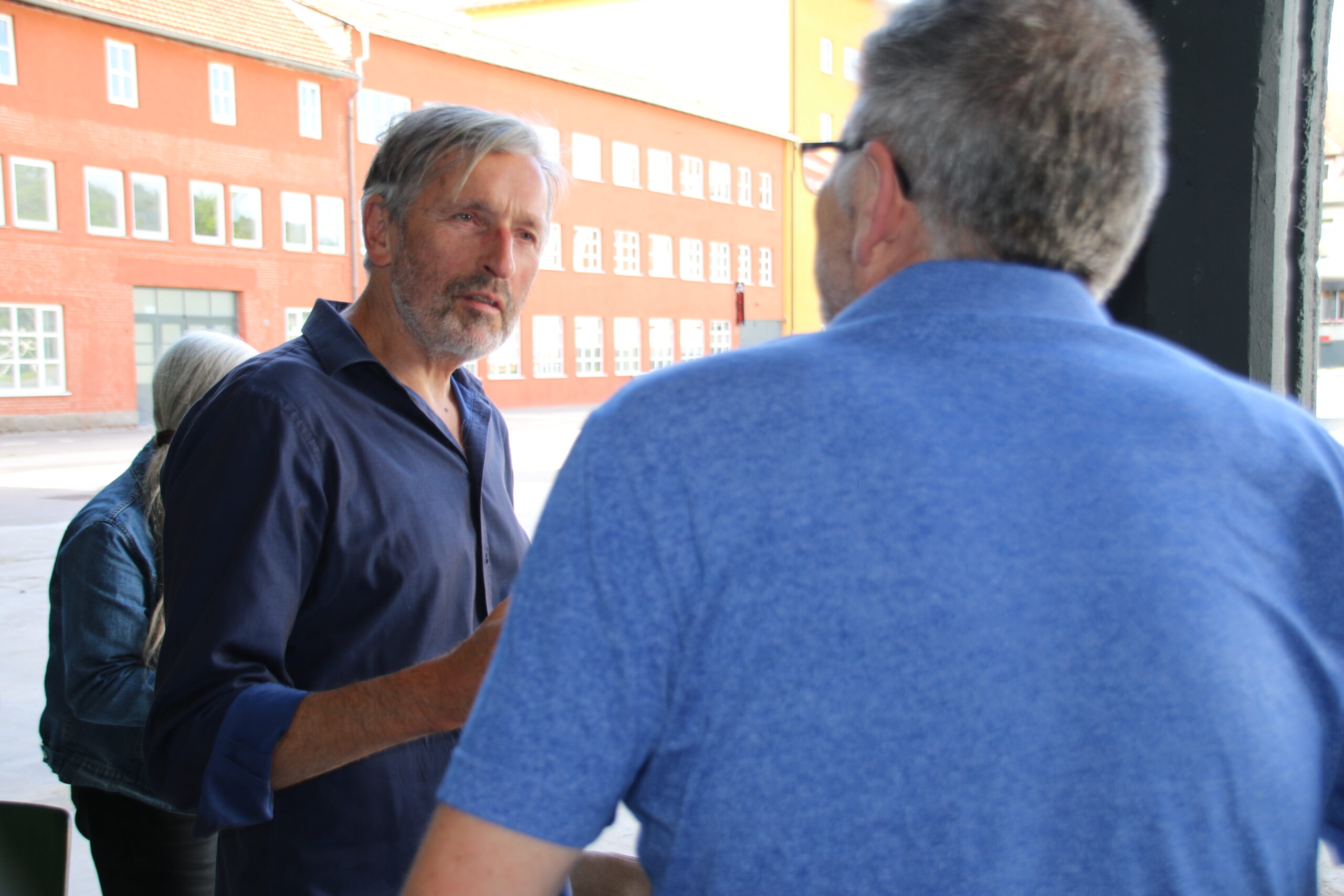 Two men are engaged in conversation outdoors near Hochschule Coburg. One man with gray hair and a beard is gesturing with his hand, while the other has short gray hair and is wearing a blue shirt. A brick building is visible in the background.