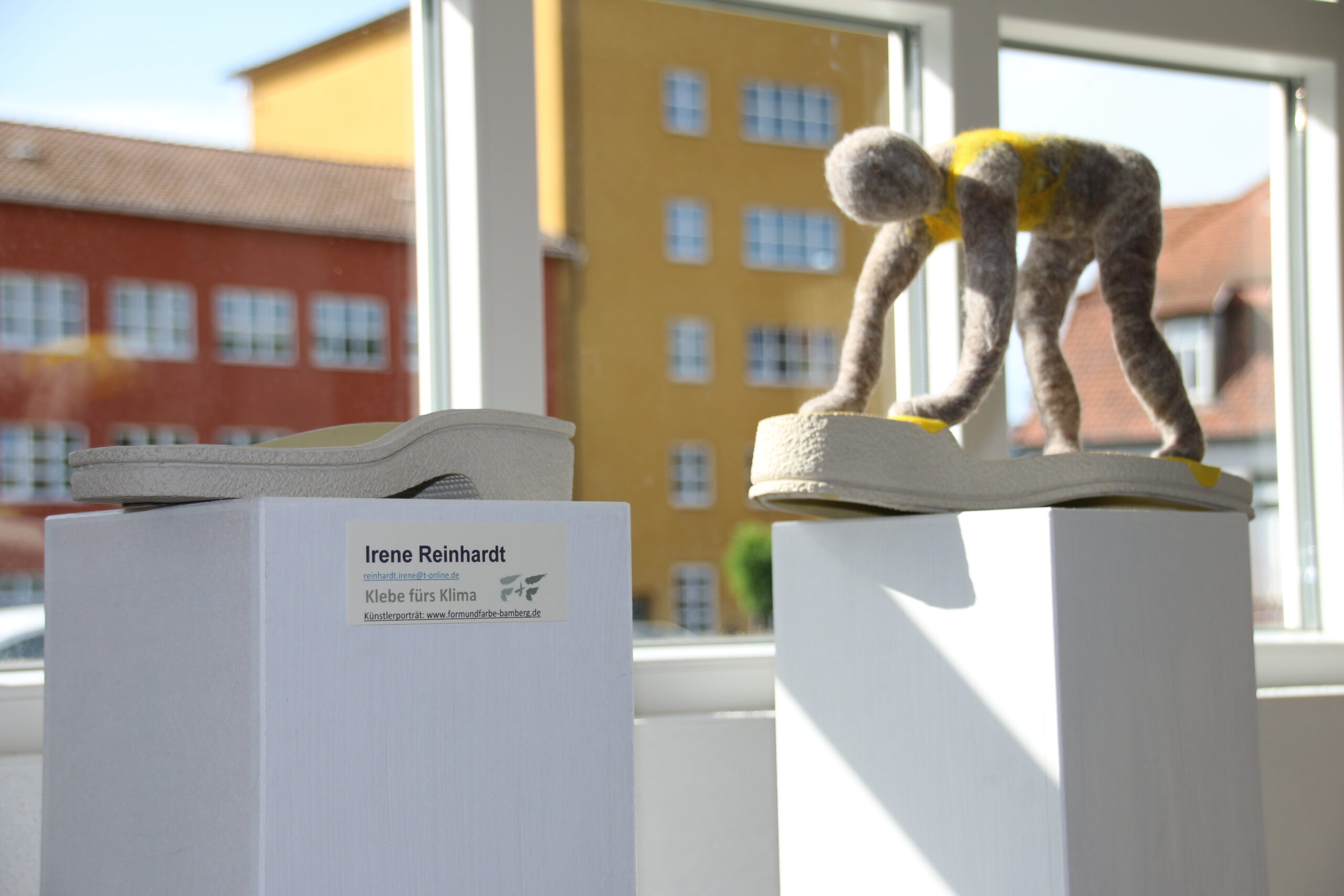 In a sunlit room at Hochschule Coburg, an art installation features two pedestal-mounted sculptures resembling wooly figures on shoe soles. The label on the left reads "Irene Reinhardt, Klebe fürs Klima," with bright buildings visible outside the window.