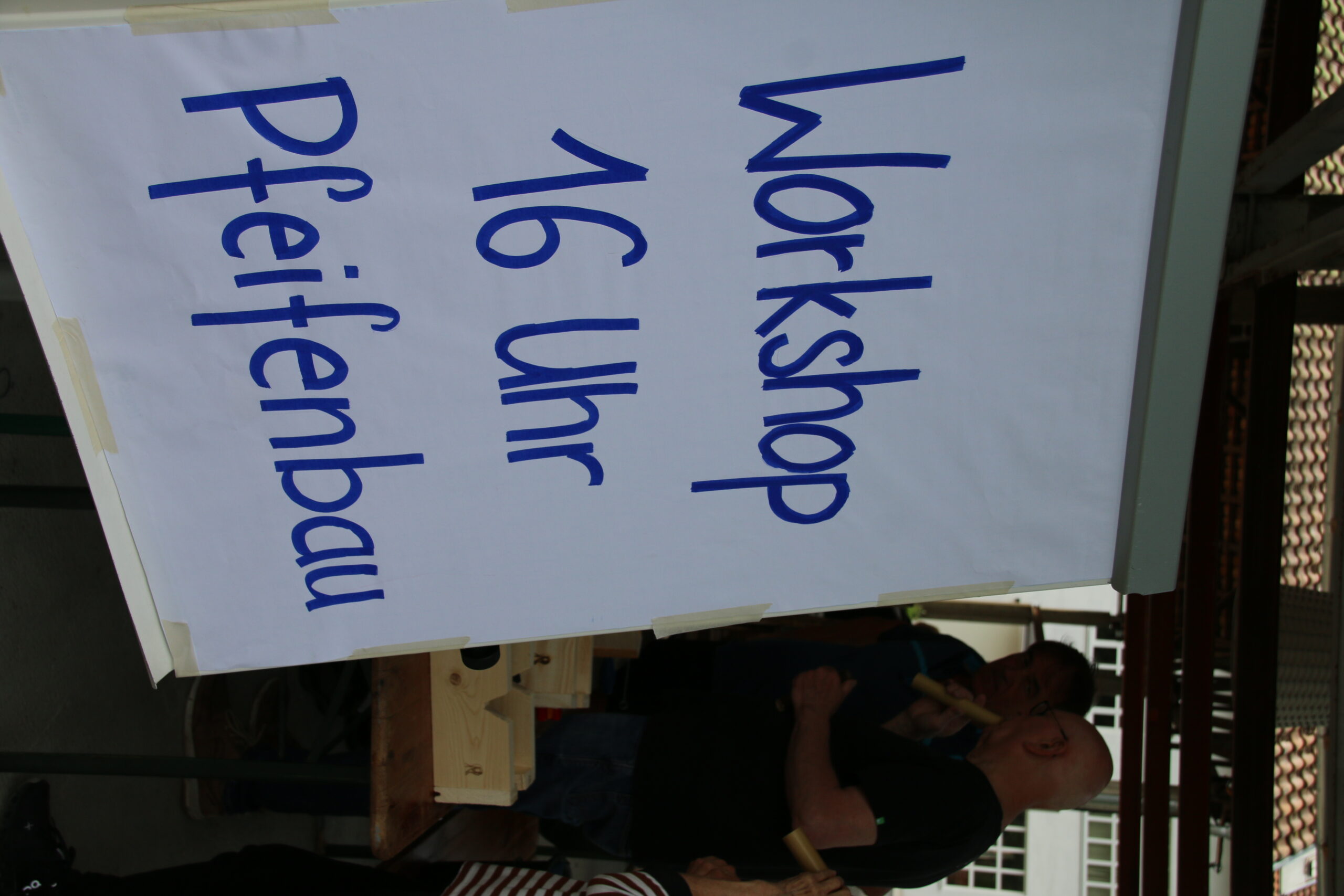 A sign with blue text reads, "Workshop 16 Uhr Pfeifenbau," near a group of people gathered around a table with wooden items. This outdoor scene unfolds under a shelter, possibly at Hochschule Coburg.