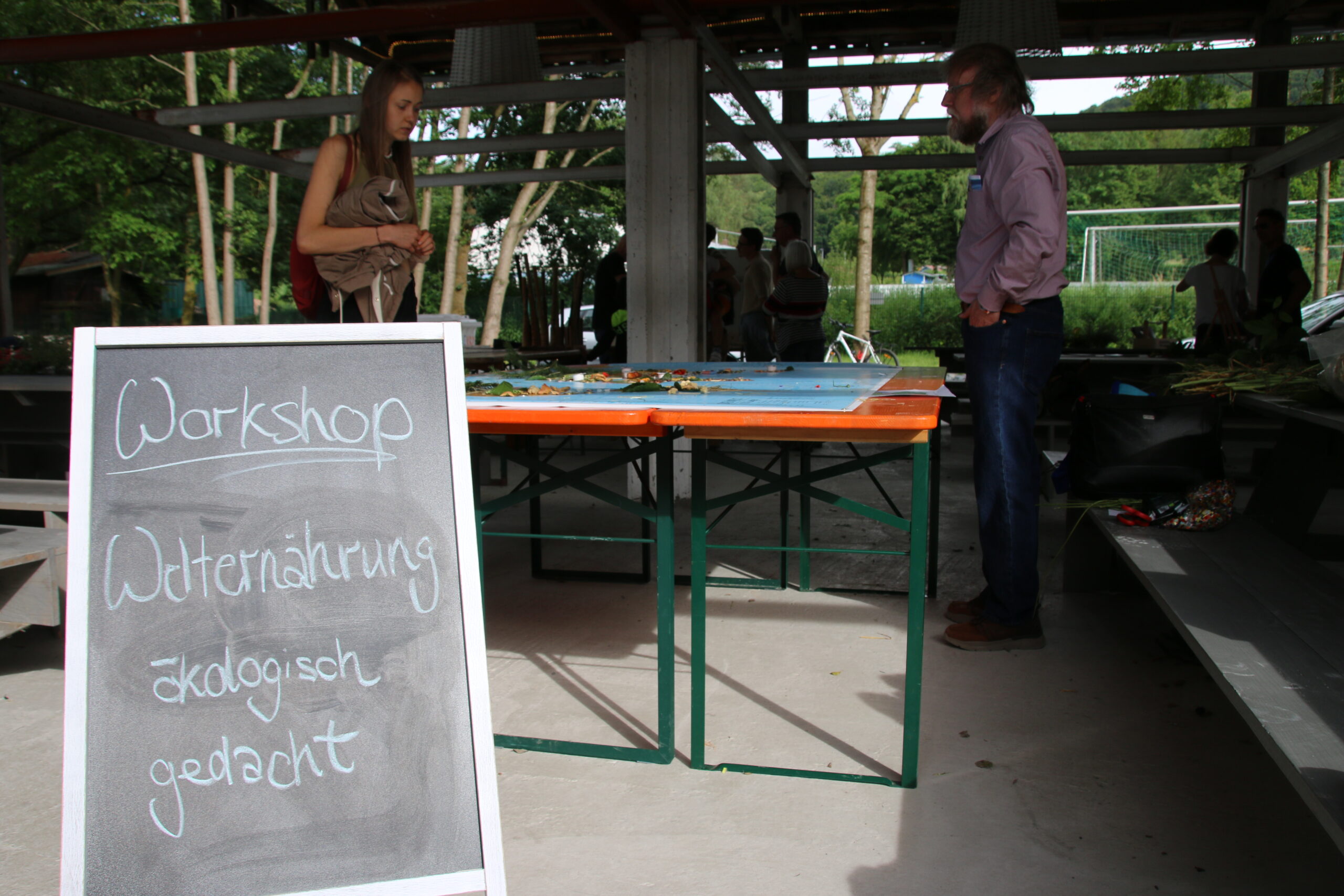 Eine Werkstattkulisse mit einer Tafel mit der Aufschrift „Workshop Wilternährung ökologisch gedacht“. Zwei Personen, möglicherweise mit der Hochschule Coburg verbunden, unterhalten sich an mit Materialien beladenen Tischen. Die Szene wird durch eine von Bäumen gesäumte, parkähnliche Umgebung im Hintergrund bereichert.