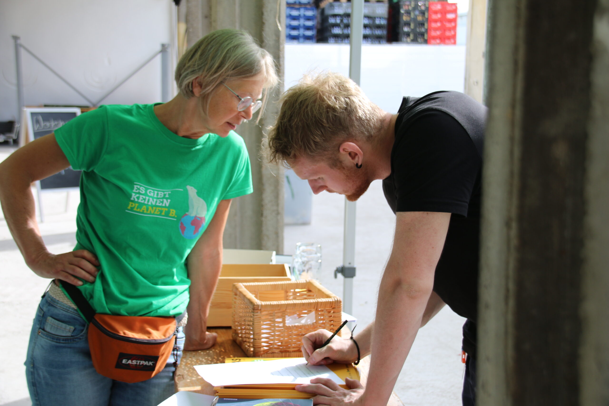 Two people engaged in conversation at a table. The person on the left, possibly a student from Hochschule Coburg, wears a green T-shirt and glasses, while the person on the right, in a black shirt, is writing on paper. A wicker basket is on the table. The setting appears casual.